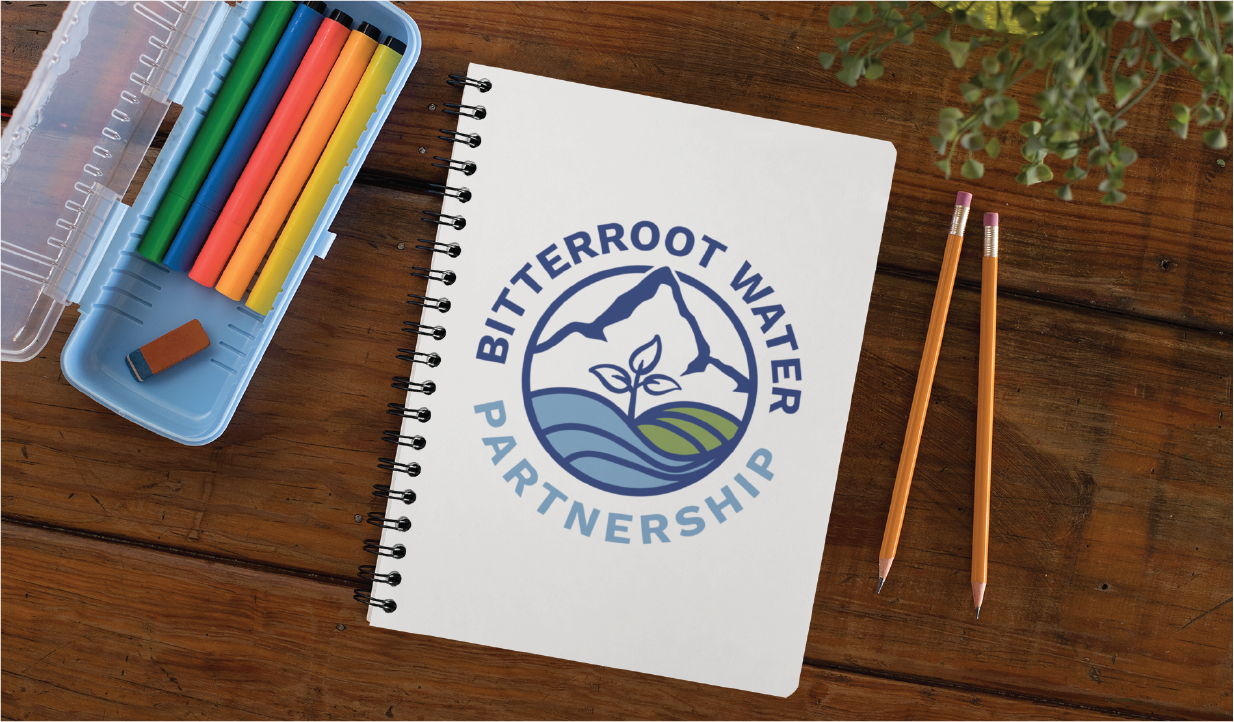 Notebook displaying the Bitterroot Water Partnership logo on a wooden desk beside colored pencils and a pencil case.