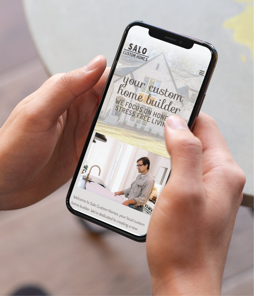 A person holds a smartphone displaying the Salo Custom Homes website, which features modern website design details about custom home building and an image of a man working in a sleek California kitchen.