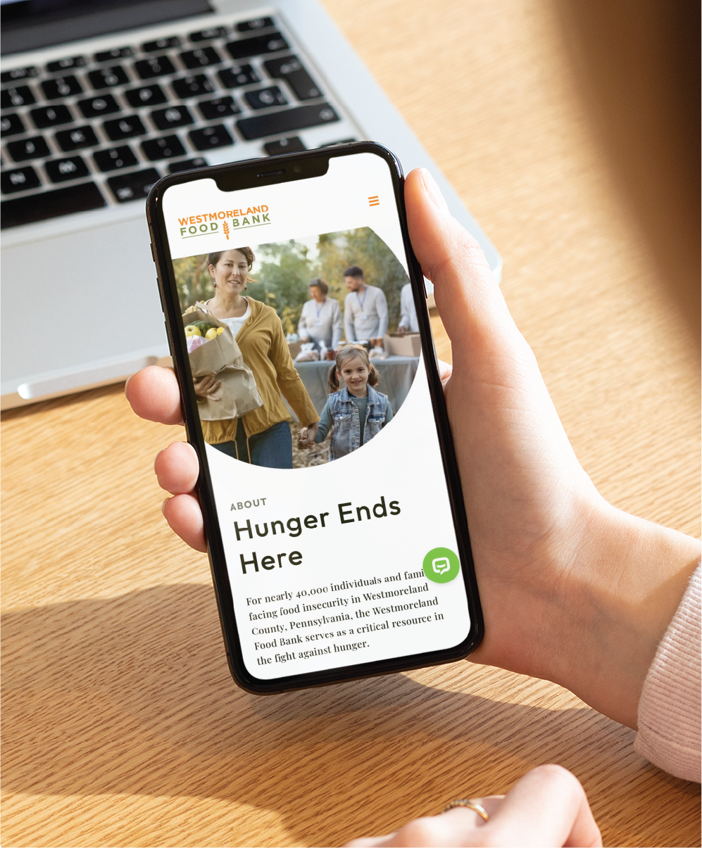 A person holds a smartphone displaying the Westmoreland Food Bank website design, featuring a smiling woman and child with groceries and the headline Hunger Ends Here on a wooden table background.