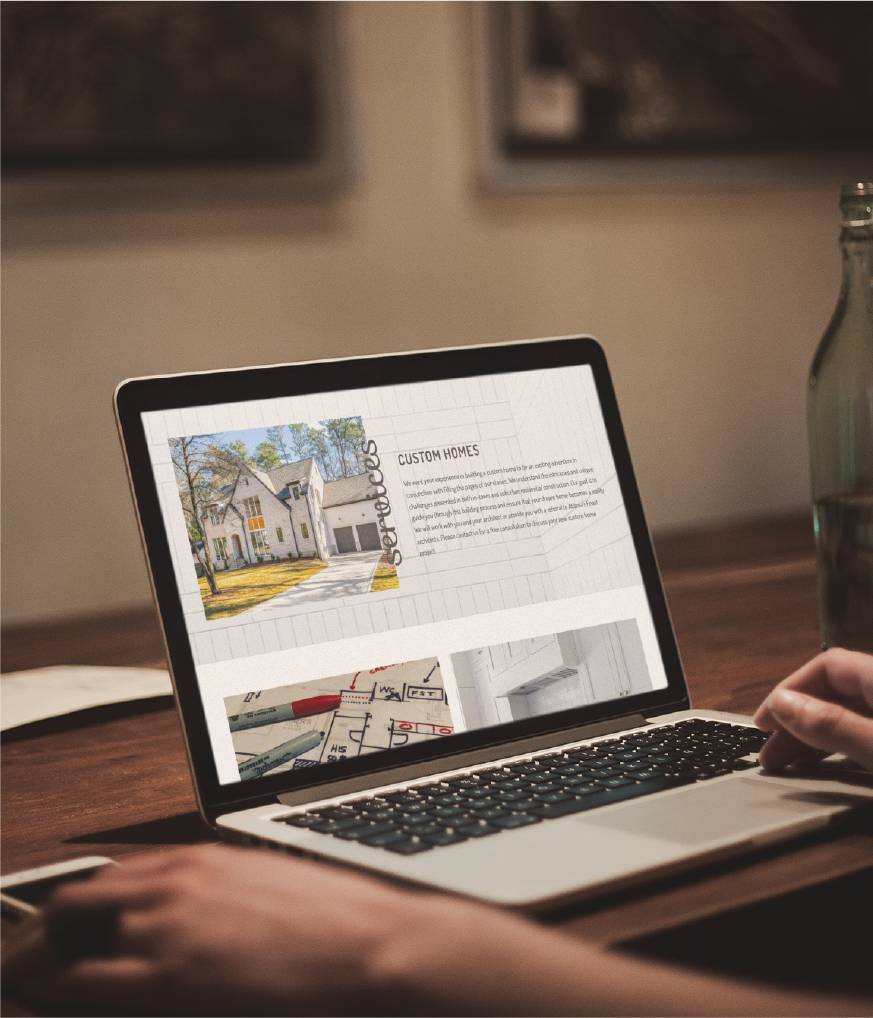A person researching Website Design for custom homes in California sits at a wooden table, browsing a site filled with house images and architectural plans, with a glass bottle nearby.