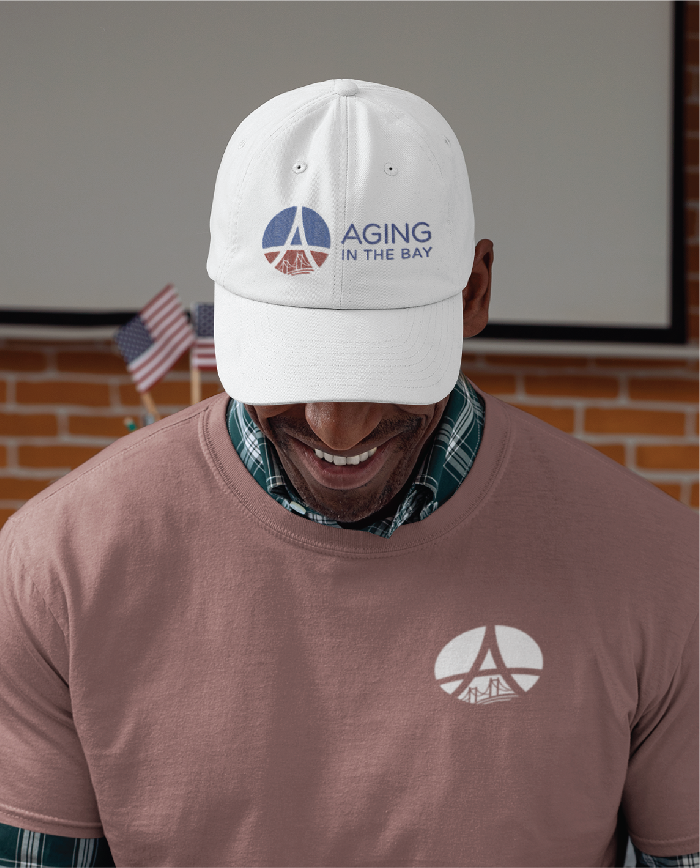 A smiling man wearing a white cap and mauve shirt, both featuring the Aging in the Bay logo design. Small American flags and a brick wall are visible in the background, adding charm to this Montana-inspired scene.