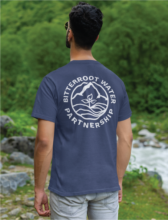 A person stands outdoors near rocks and greenery, facing away. They wear a blue T-shirt with a circular logo design and the text Bitterroot Water Partnership on the back, blending in with the calm beauty of a nature preserve.