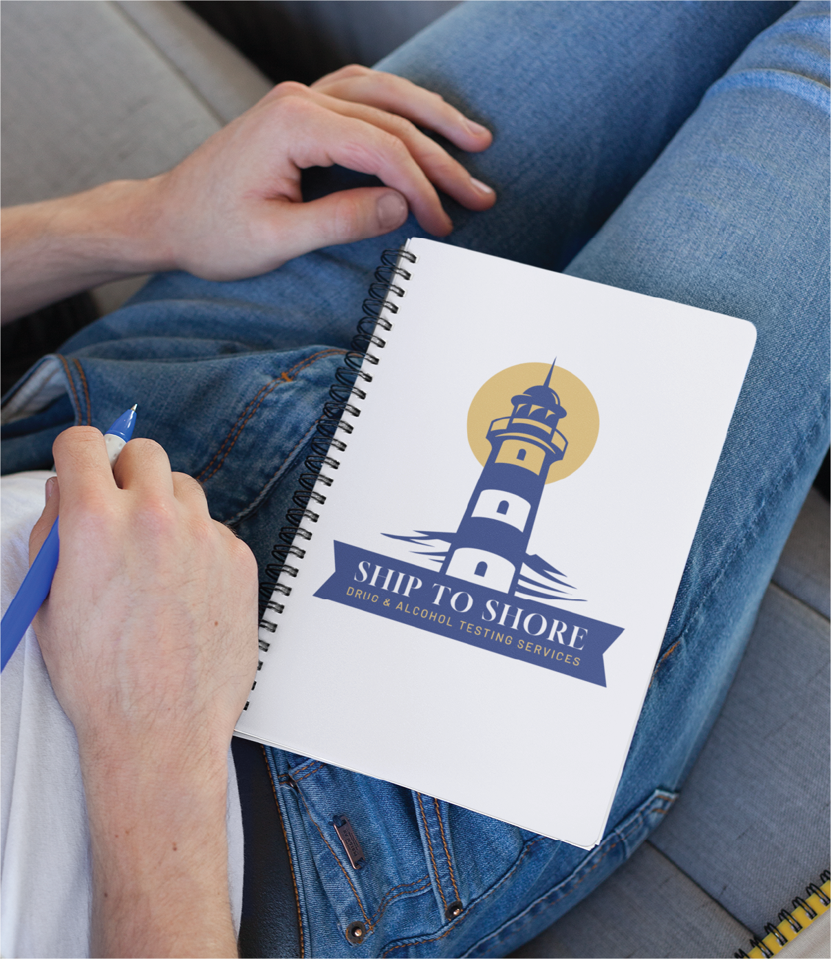 A person in blue jeans holds a pen and a spiral notebook featuring a Professional Logo Design—a lighthouse with the text Ship to Shore: Drug & Alcohol Testing Services—on the cover.