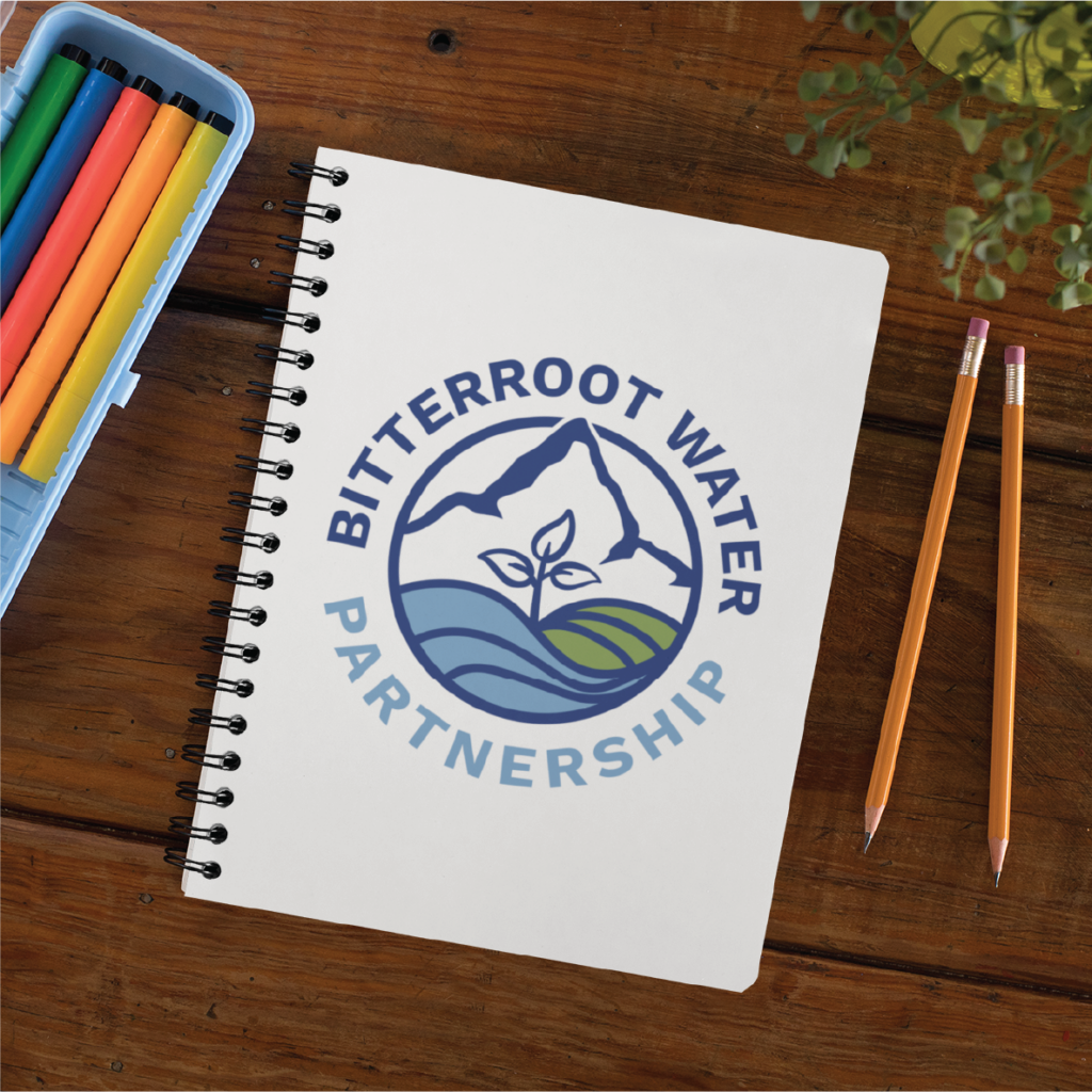 A spiral notebook with the Bitterroot Water Partnership logo design, featuring a mountain, river, and plant, sits on a wooden desk with colored markers, two pencils, and a green plant in the corner—evoking the feel of a nature preserve.