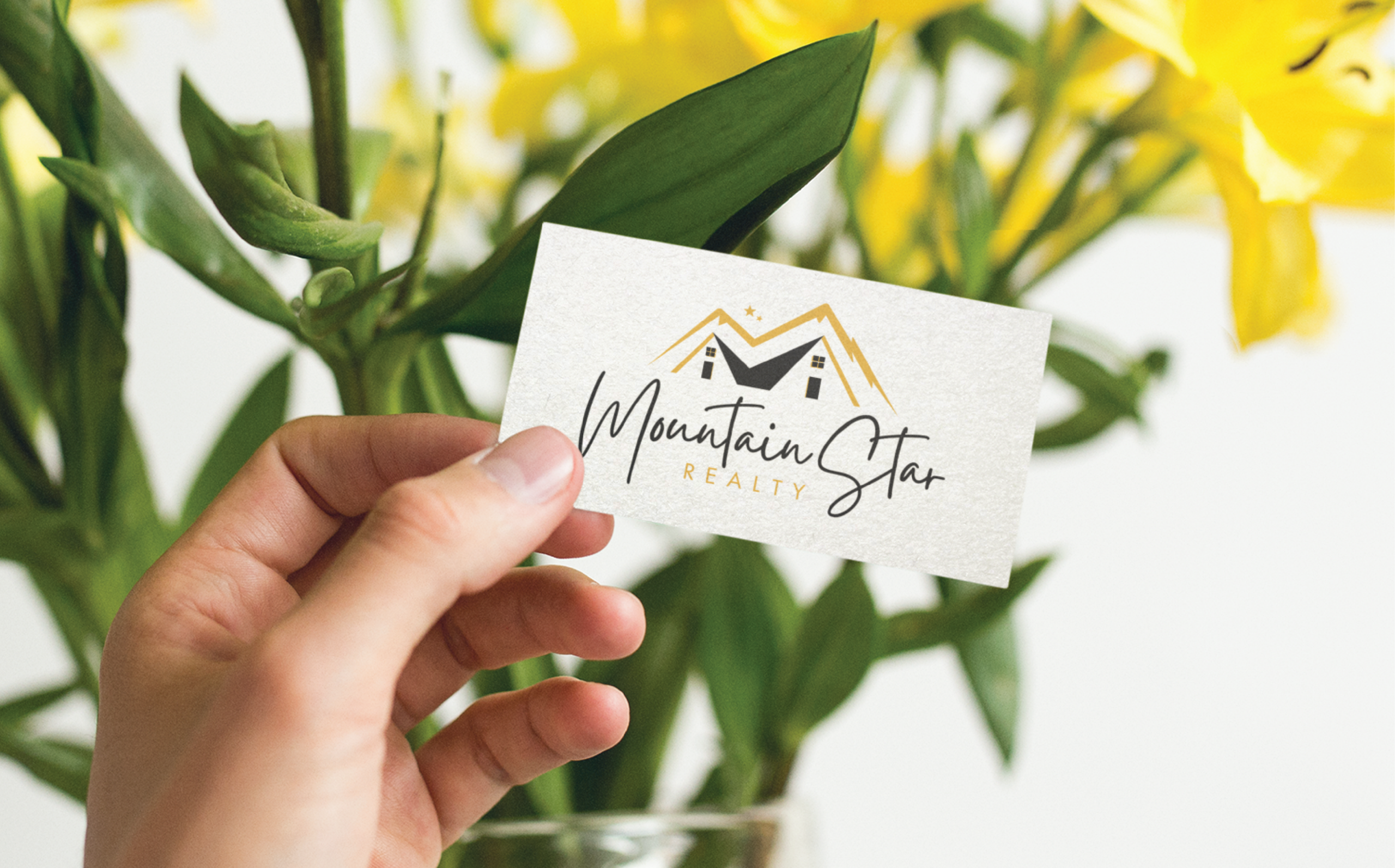 print and grpahic design for realty business | Moonlit Media LLC A hand holding a business card with the logo and text Mountain Star Realty in front of yellow flowers and green leaves. The card features a stylized mountain, house design, and reflects professional stationery design for Michigan clients.