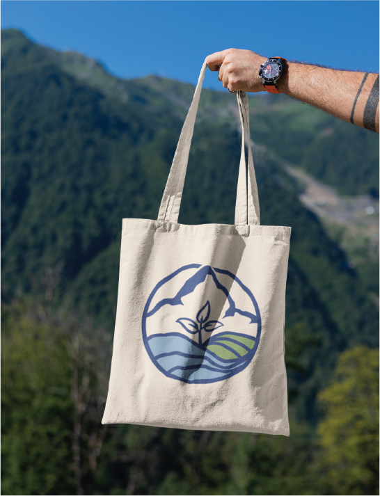 Beige tote bag with a circular nature logo held up against a mountain landscape.