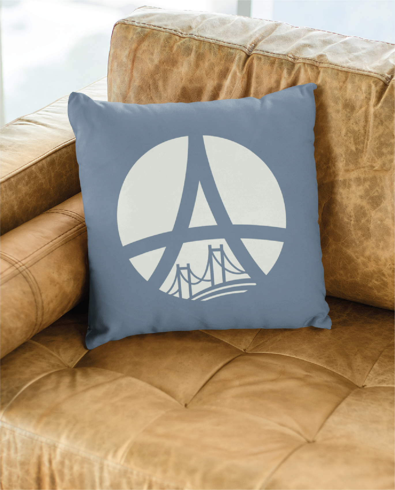 A blue pillow with a white graphic design featuring the Eiffel Tower above a bridge, inspired by logo design, placed on a tan leather couch near a window.