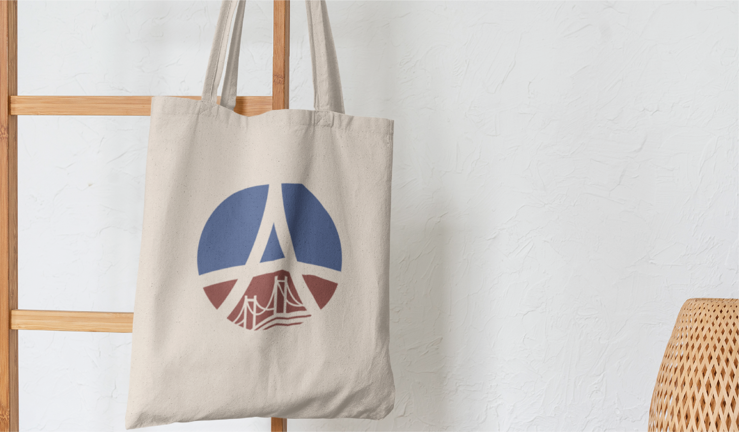 A beige tote bag hangs on a wooden ladder, featuring a striking logo design with blue and red geometric shapes resembling a stylized letter A and a bridge. The background is a white textured wall, evoking the spirit of Montana’s scenic nature preserves.