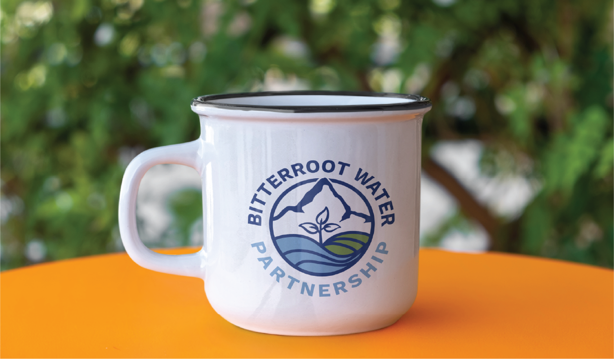A white metal mug with a black rim sits on an orange surface. The mug displays the Bitterroot Water Partnership logo, designed through custom logo creation, featuring a mountain, water, and leaf graphic inspired by Montana’s nature preserves. Green foliage is blurred in the background.