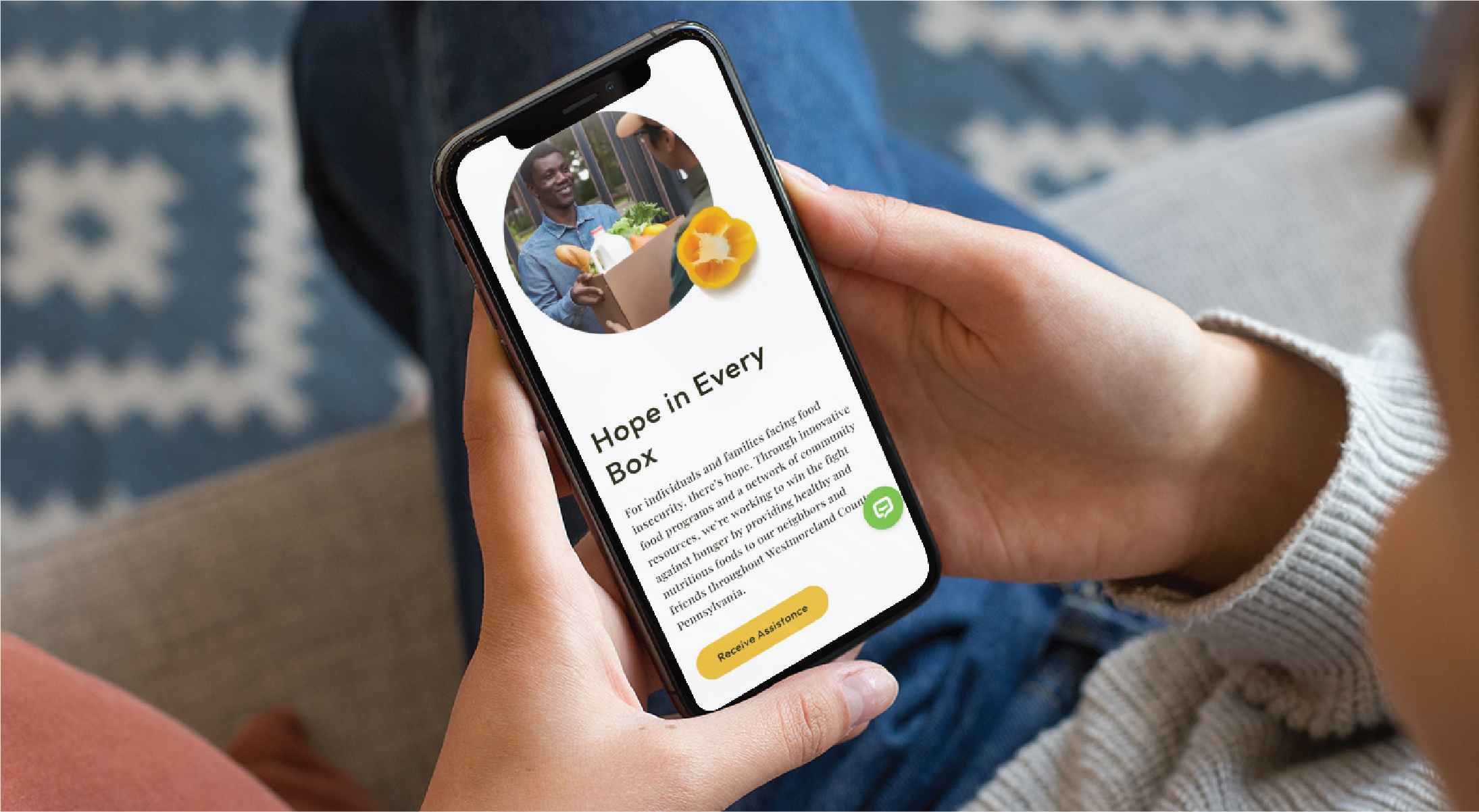A person in California holds a smartphone displaying a website design titled “Hope in Every Box,” featuring smiling faces, a yellow pepper, and buttons at the bottom. The phone rests casually on their lap, creating a relaxed wellness practice vibe.