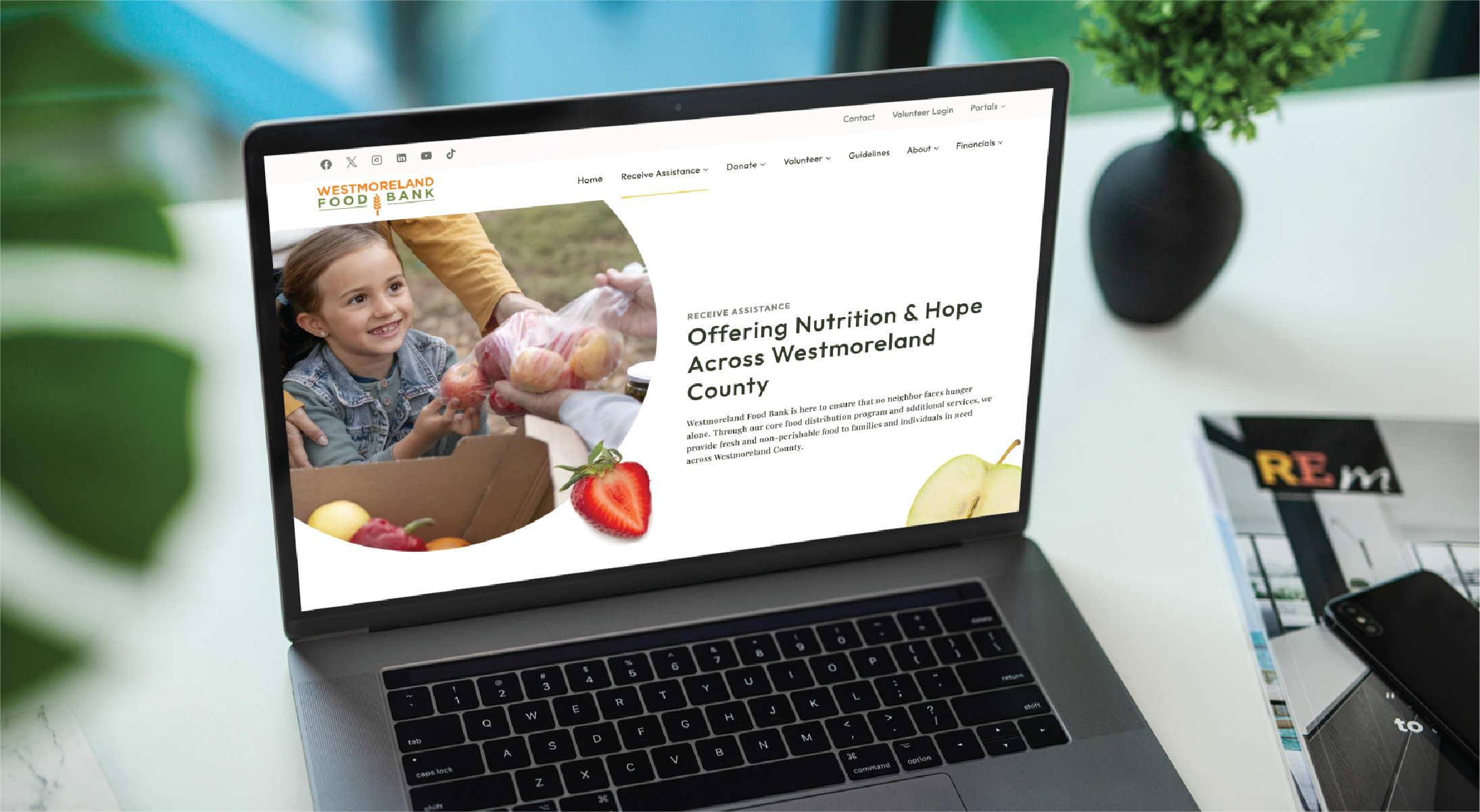 A laptop showcases the Westmoreland Food Bank website design, featuring a smiling child holding apples and text about offering nutrition and hope. The laptop sits on a desk with a plant, magazine, and smartphone nearby.