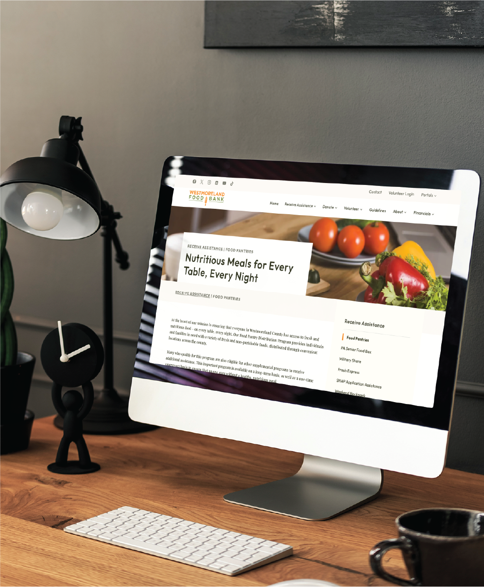 A desktop computer on a wooden desk displays a food pantry website, showcasing thoughtful website design. Nearby are a lamp, clock, keyboard, mouse, potted plant, and a small black figurine sitting by the monitor.