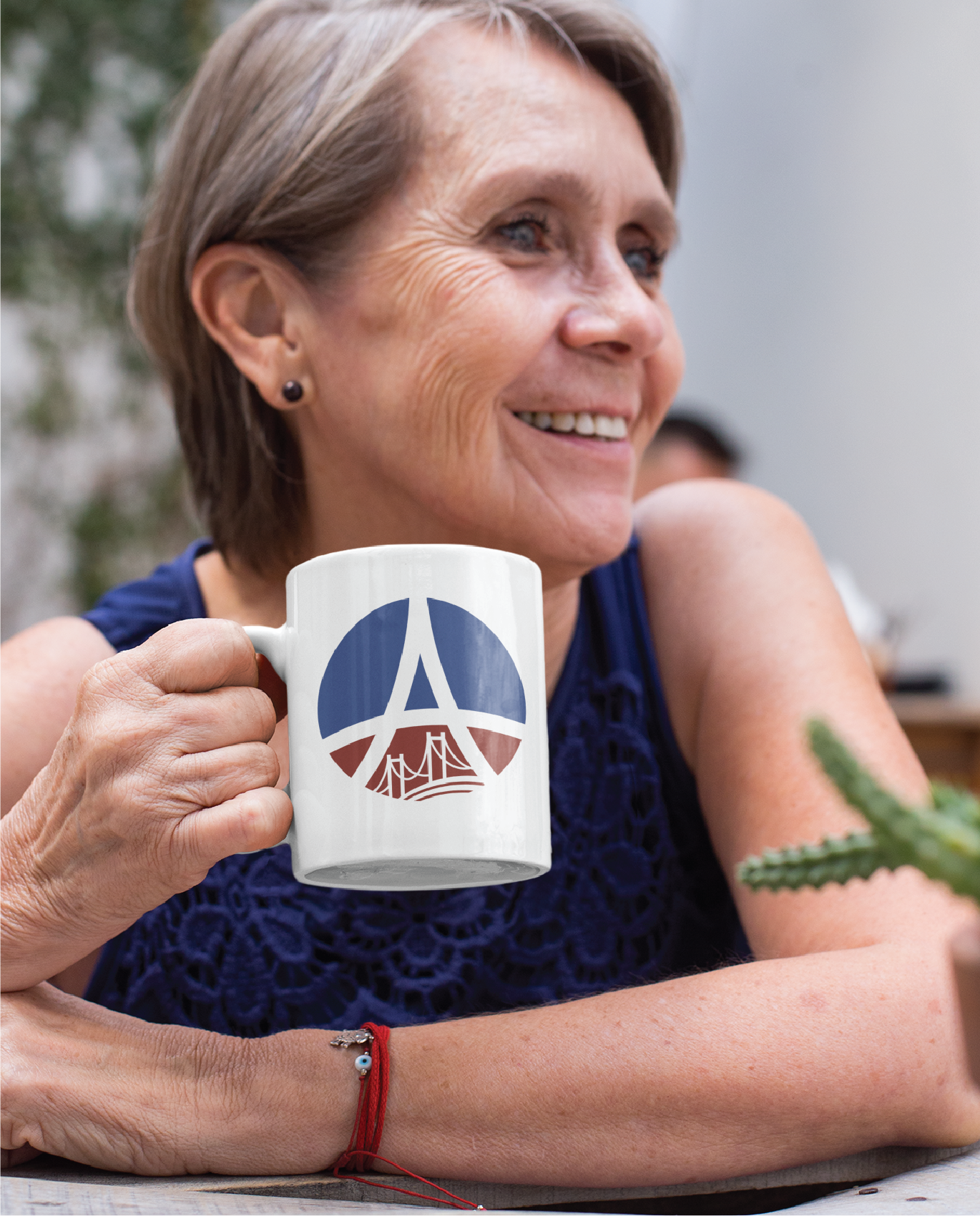 Smiling older woman with short gray hair holds a white mug featuring a stylized Montana logo design in blue, white, and red. She’s seated at a table, wearing a navy sleeveless top and red string bracelet. A small potted plant is in the foreground.
