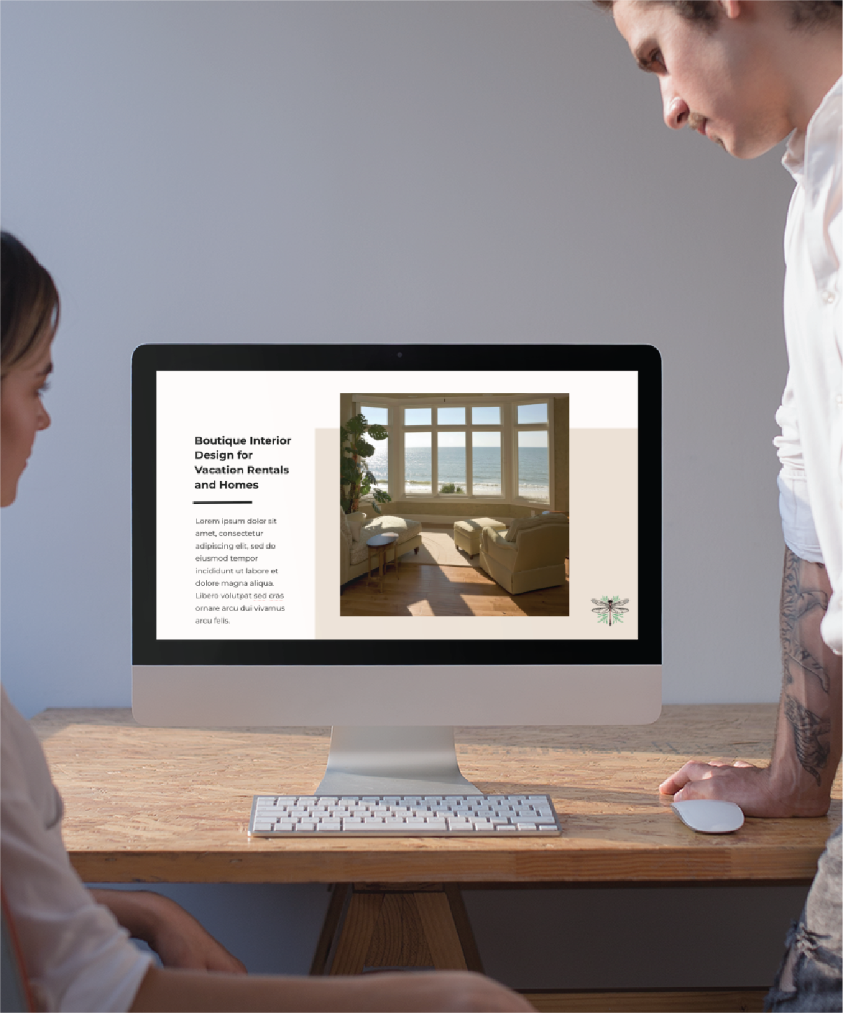 Two people stand at a desk, looking at a computer screen displaying an Interior Designer's webpage about boutique interior design for vacation rentals and homes in Florida, featuring social graphics and a photo of a bright living room with large windows.
