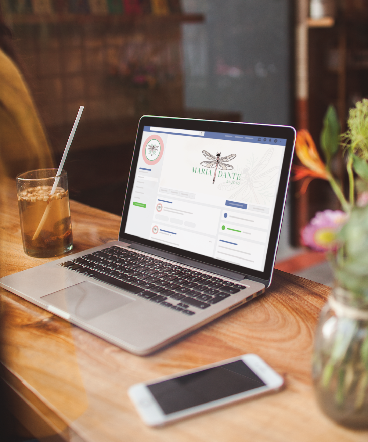 A laptop displaying a profile page with a dragonfly logo is on a wooden table next to a glass of iced drink, smartphone, and flowers in a cozy café—perfect inspiration for an Interior Designer in Florida working on new Social Graphics.