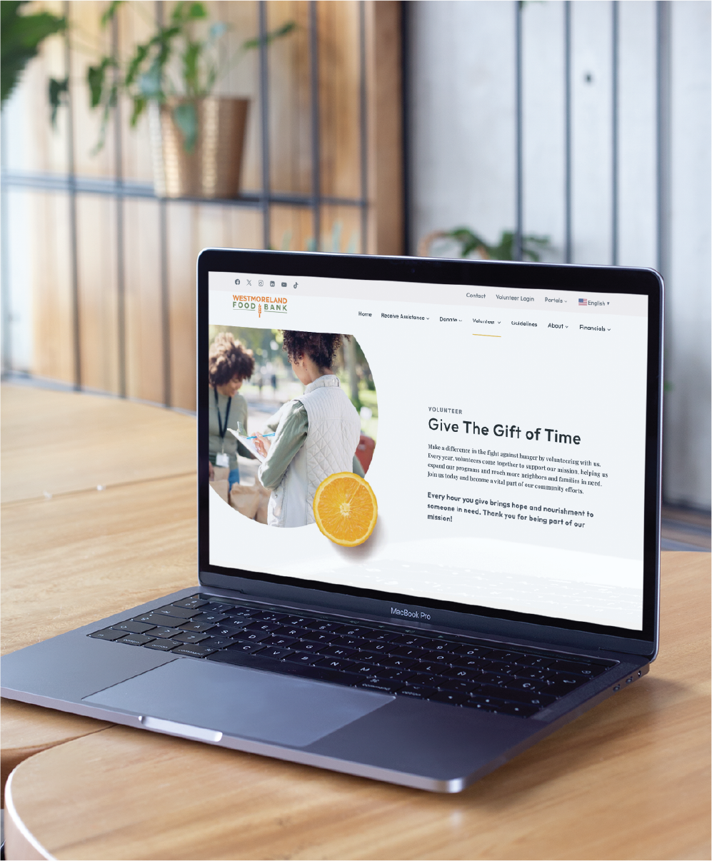 A laptop on a wooden table displays a website about volunteering, highlighting California community efforts. The screen shows two people volunteering, text about giving time, and a slice of orange in a modern, bright room with plants—an inviting website design.