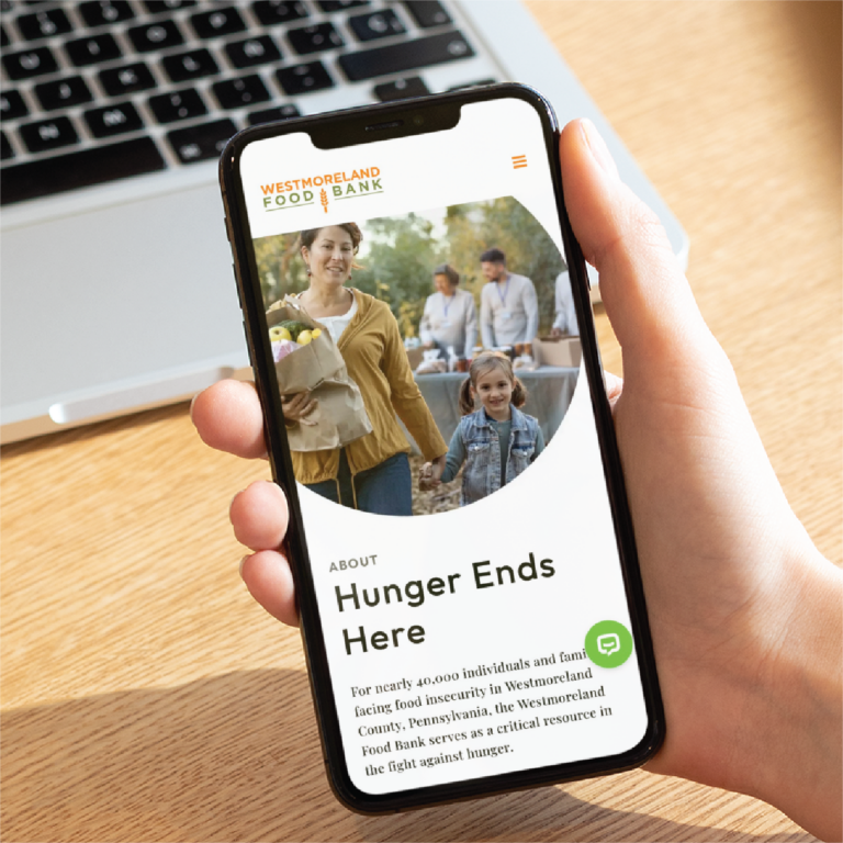 A hand holds a smartphone displaying the Westmoreland Food Bank website with a modern website design, featuring an image of a woman and child with groceries. A laptop is blurred in the background on a light wooden surface.