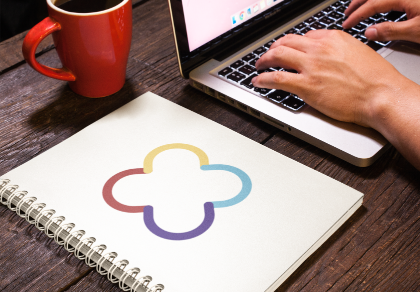 A person types on a laptop at a wooden table with a red coffee mug and a spiral-bound notebook featuring an AI-generated logo in a colorful, abstract four-lobed design on the cover.