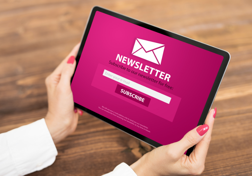 A person holding a tablet displaying a pink newsletter sign-up screen with an envelope icon, email input field, and Subscribe button on a wooden table—highlighting small business privacy during online engagement.