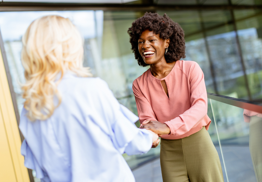 Two women are standing and smiling while shaking hands on an outdoor balcony, discussing privacy basics for small business. One has curly hair and a pink blouse; the other sports blonde hair and a light blue shirt. The atmosphere appears friendly and professional.