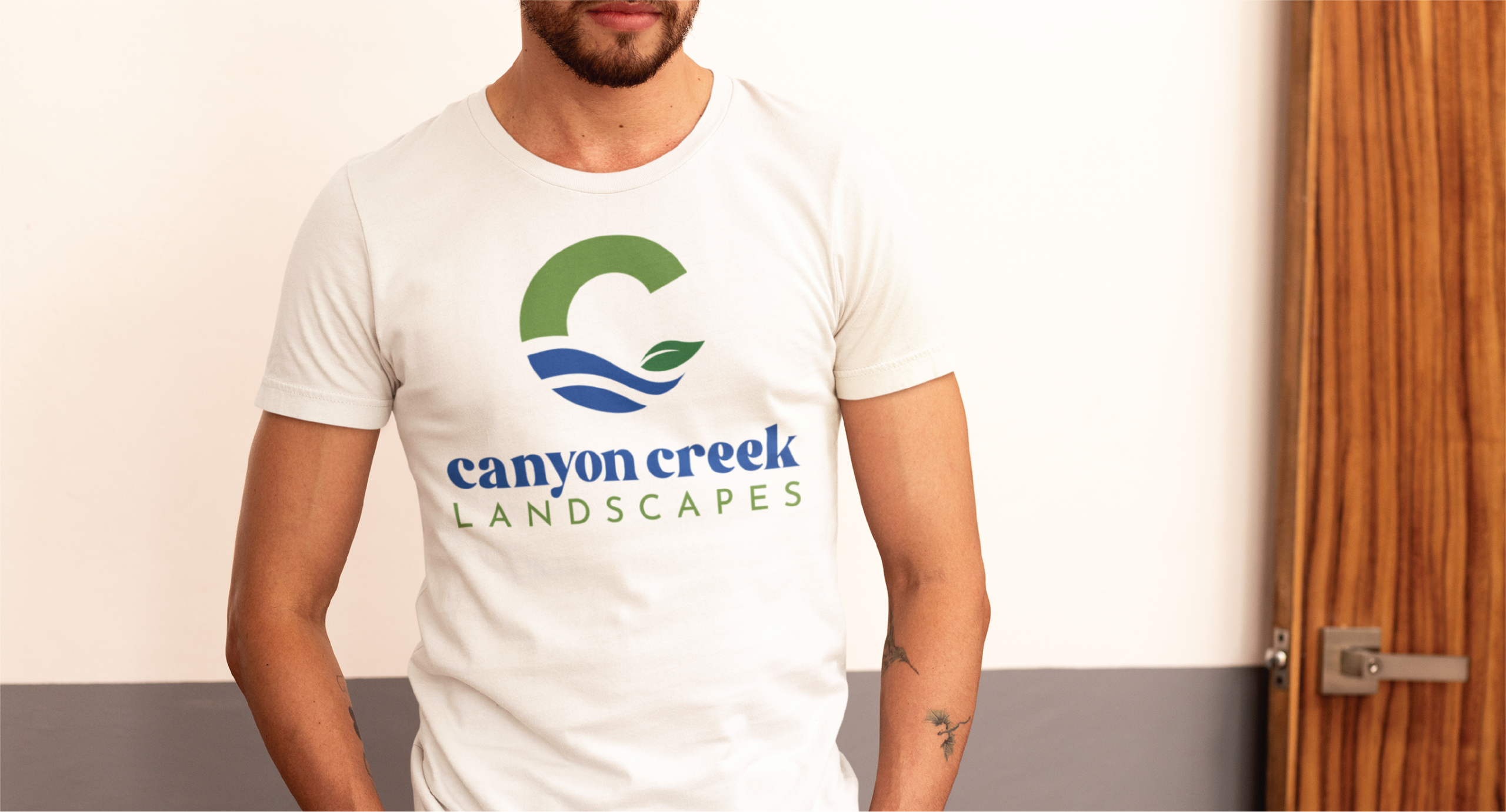 A person wearing a white t-shirt with the Canyon Creek Landscapes brand design—featuring a green C with a leaf and blue water waves—stands indoors near a wooden door, representing this New Jersey business with style.