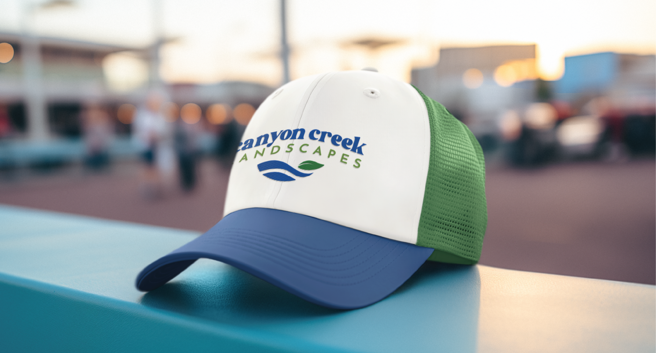 A green and white trucker hat with a blue brim rests on a teal surface. Featuring the Canyon Creek Landscapes logo and leaf-water graphics, this hat highlights expert brand design. The softly blurred background hints at New Jersey’s outdoor charm.