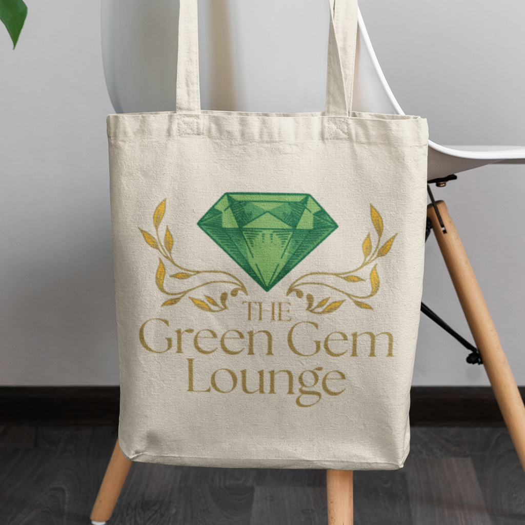 A beige tote bag hangs on a white chair. The bag features a large green gem logo design above gold decorative branches and the text “The Green Gem Lounge.” The scene is set against a dark wooden floor.