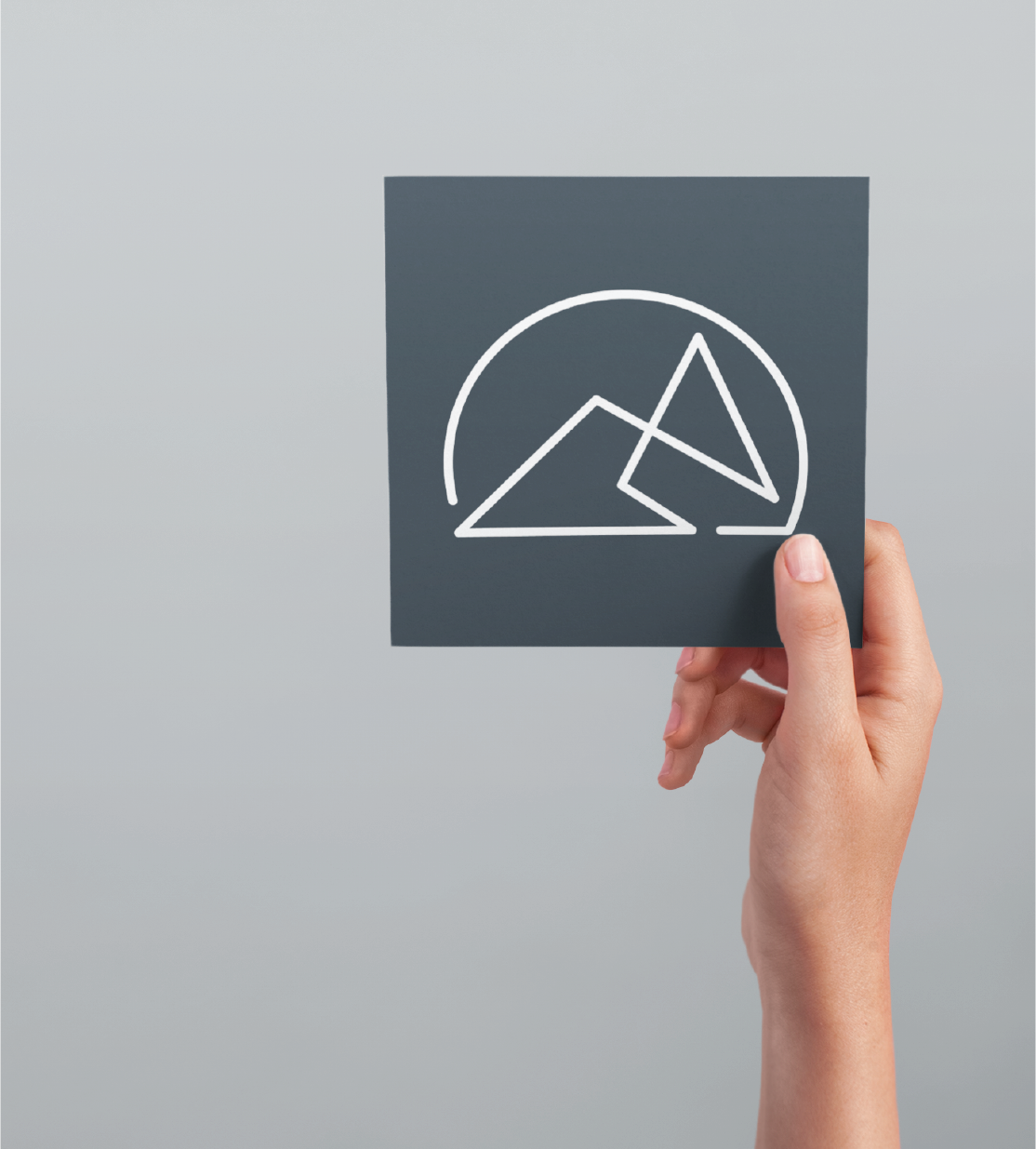 A hand holding a sleek business card with a minimalist white line drawing of two mountains and a semicircle, representing the sun or moon, against a plain light gray background—perfect inspiration for business card design in Michigan.