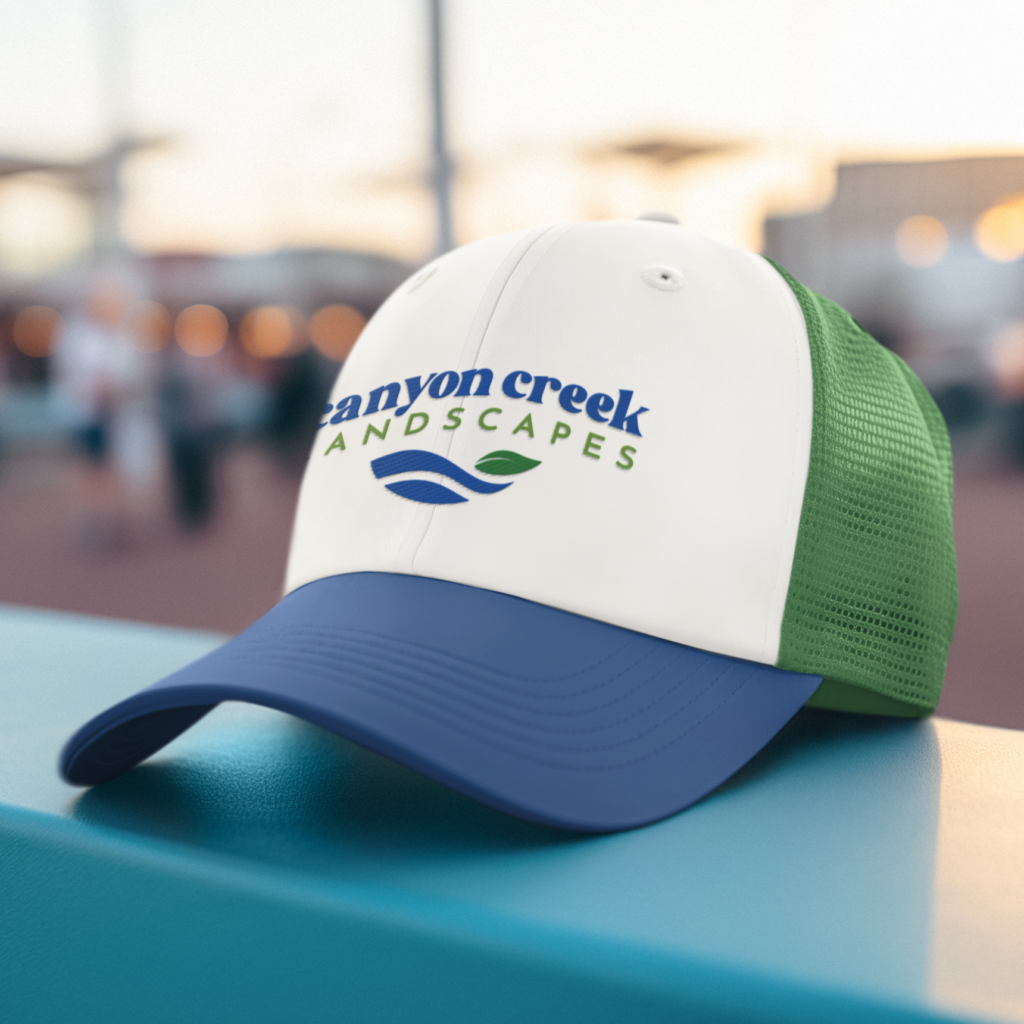 A white and green trucker hat with a blue bill features the Canyon Creek Landscapes logo and a blue and green leaf design, showcasing standout brand design, resting on a turquoise surface outdoors.