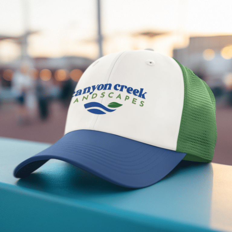A white and green trucker hat with a blue bill features the Canyon Creek Landscapes logo and a blue and green leaf design, showcasing standout brand design, resting on a turquoise surface outdoors.