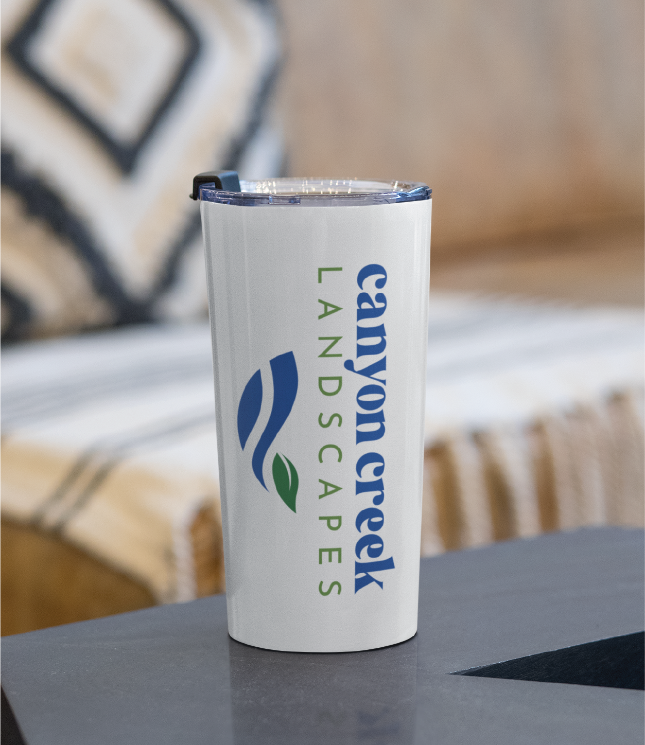 A white tumbler with a clear lid sits on a table. The tumbler showcases the Canyon Creek Landscapes logo in blue and green, highlighting thoughtful brand design. In the background, a blurred couch with patterned pillows completes the New Jersey setting.