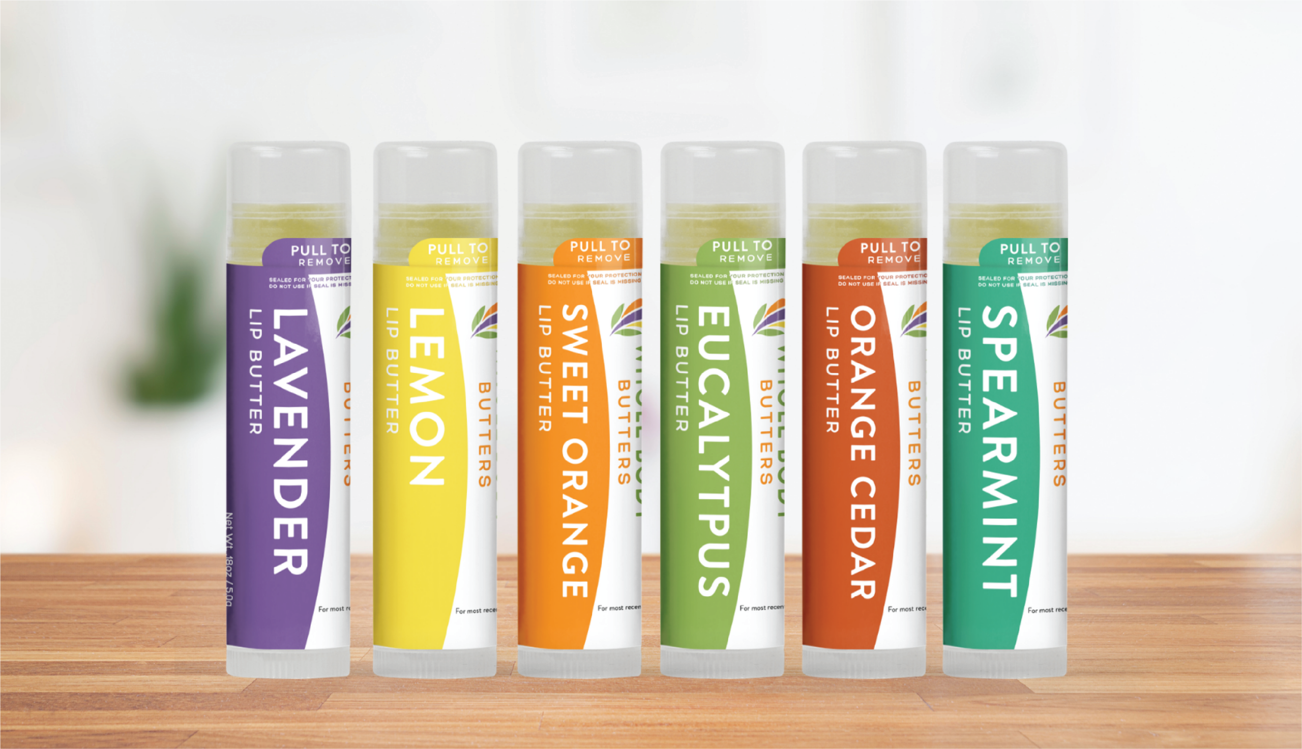 Six lip butter tubes are lined up on a wooden surface, each labeled with a different flavor. Inspired by Colorado’s natural beauty and healing arts, the vibrant packaging features thoughtful stationery design. The background is softly blurred.
