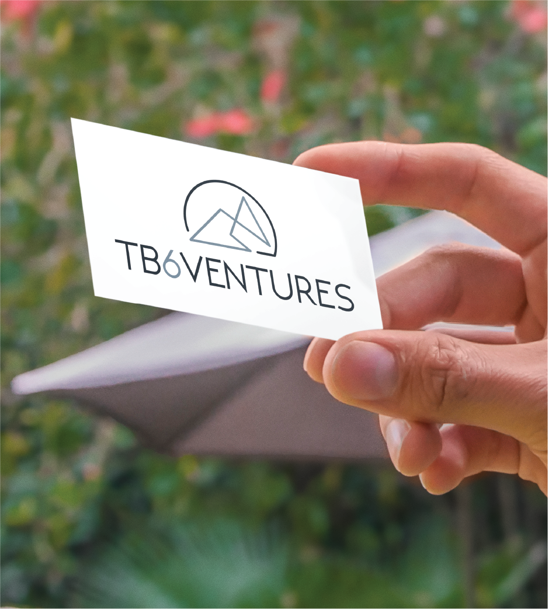 A hand holds a business card displaying the TB6Ventures logo, known for its geometric mountain design above the company name. This standout business card design reflects the quality of Michigan’s top business service providers, with blurred greenery in the background.