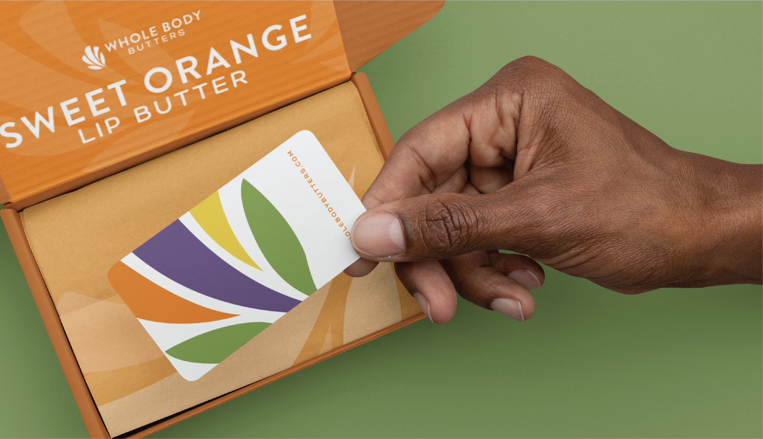 A hand holds a colorful card above an open orange box labeled Whole Body Butters Sweet Orange Lip Butter on a green surface, showcasing creative stationery design inspired by the healing arts of Colorado.