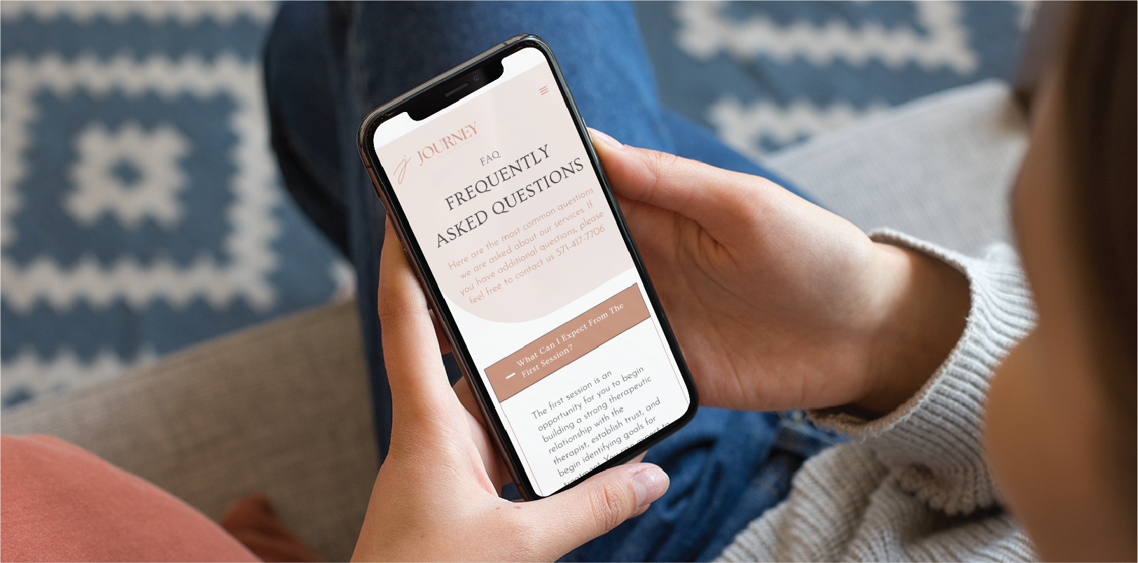 A person holds a smartphone displaying a Frequently Asked Questions webpage about Custom Web Design, with text on a beige and pink background. Seated on a couch in Georgia, they wear jeans, with a patterned rug visible in the background.
