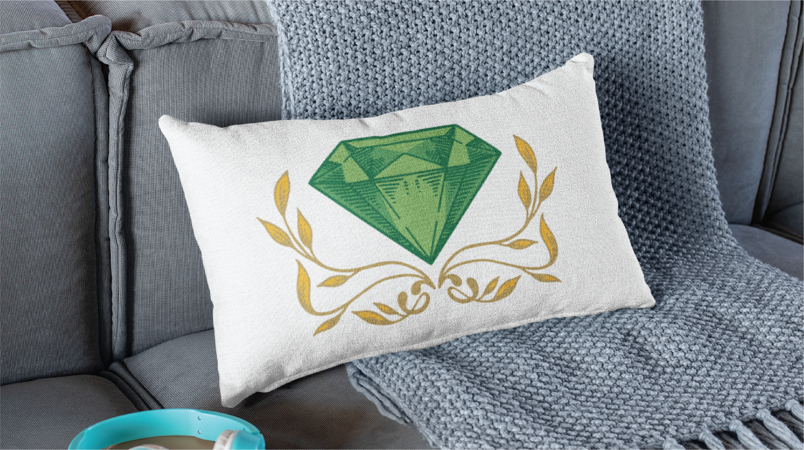 A white rectangular pillow featuring a large green diamond and gold decorative branches—a perfect touch for any Pennsylvania landscaper's lounge—rests on a gray sofa next to a textured gray blanket. A cup on a saucer sits nearby.