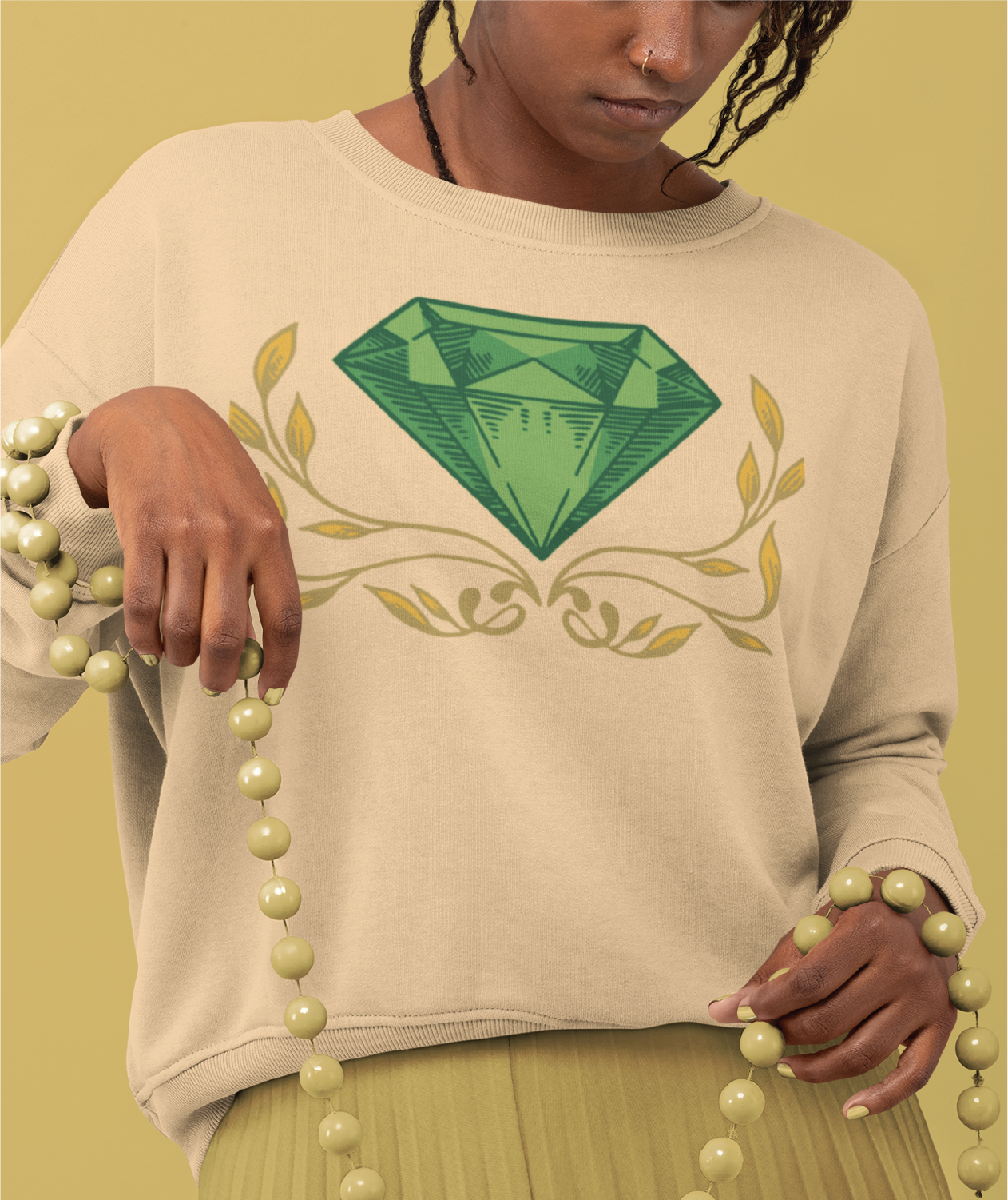 A person wearing a beige sweater with a large green diamond and decorative leaves design, reminiscent of a landscaper’s logo design, holds light green beaded necklaces against a matching beige background.