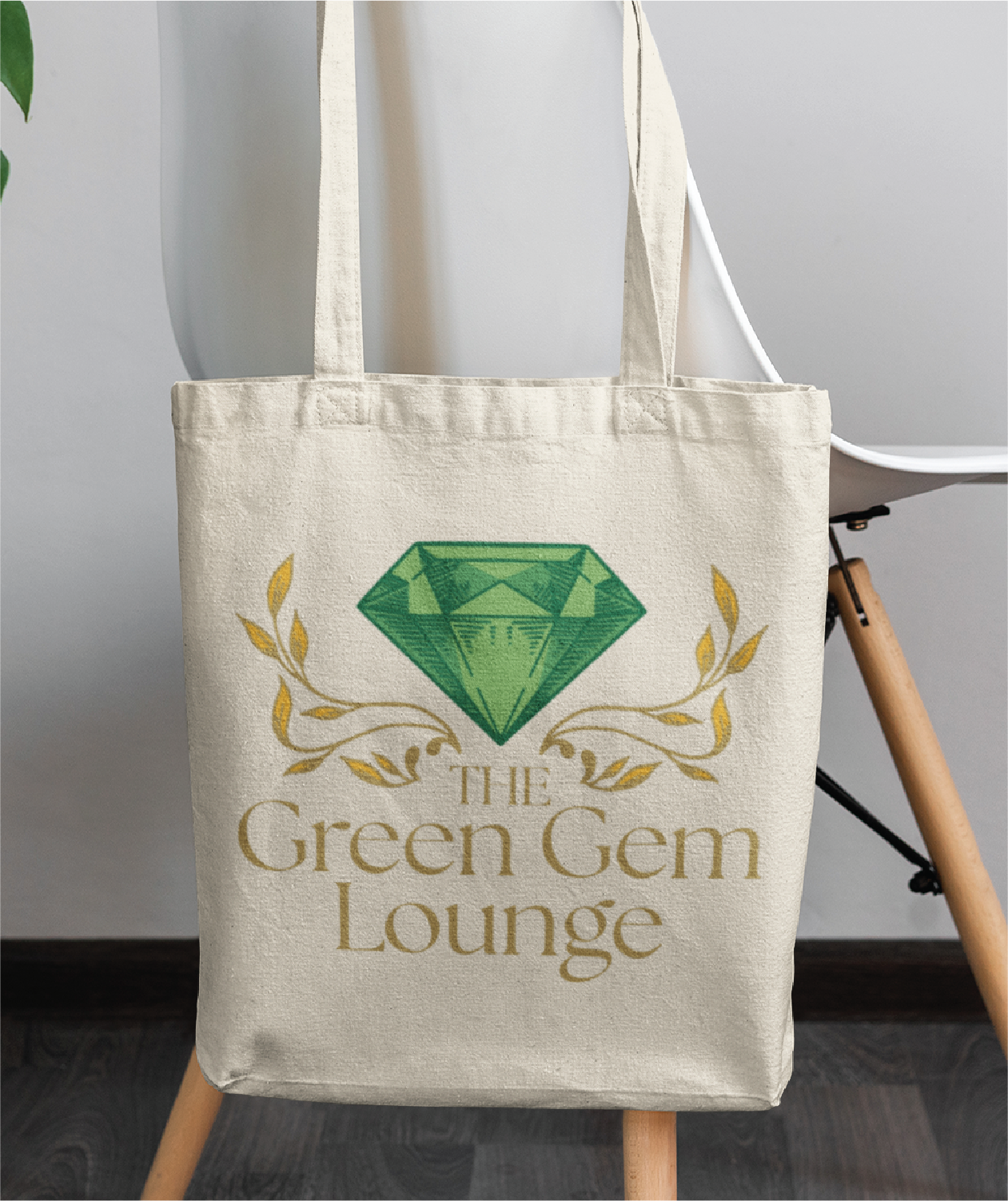 A beige tote bag hangs on a white chair. Showcasing a large green gemstone, golden leaf accents, and the text THE Green Gem Lounge below the gem, it’s perfect for any Pennsylvania landscaper seeking stylish logo design inspiration.