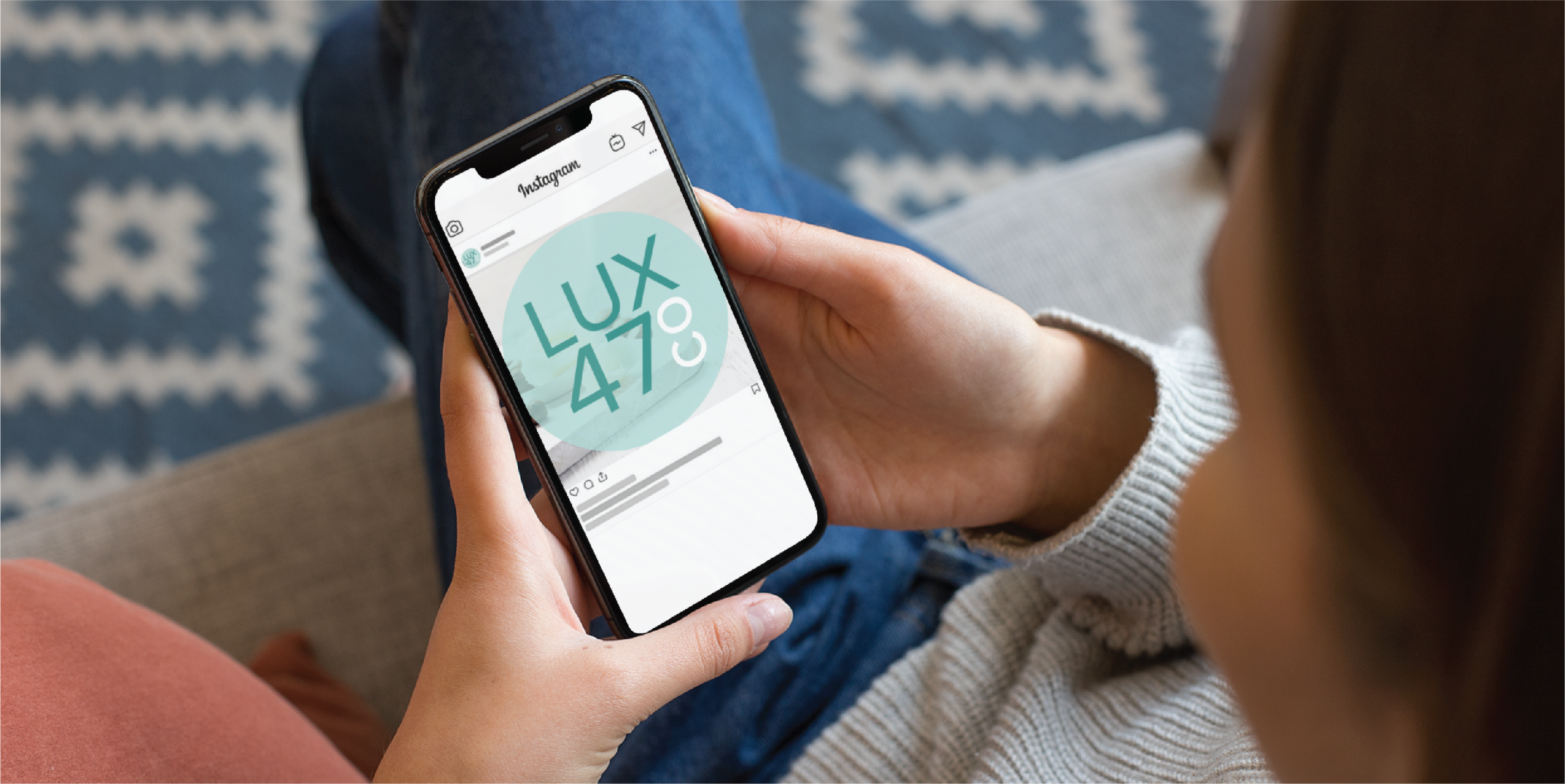 A person holding a smartphone and looking at an Instagram post displaying a circular Social Graphics design with the text LUX 47° on the screen. The person is sitting on a couch with a patterned rug in the background.