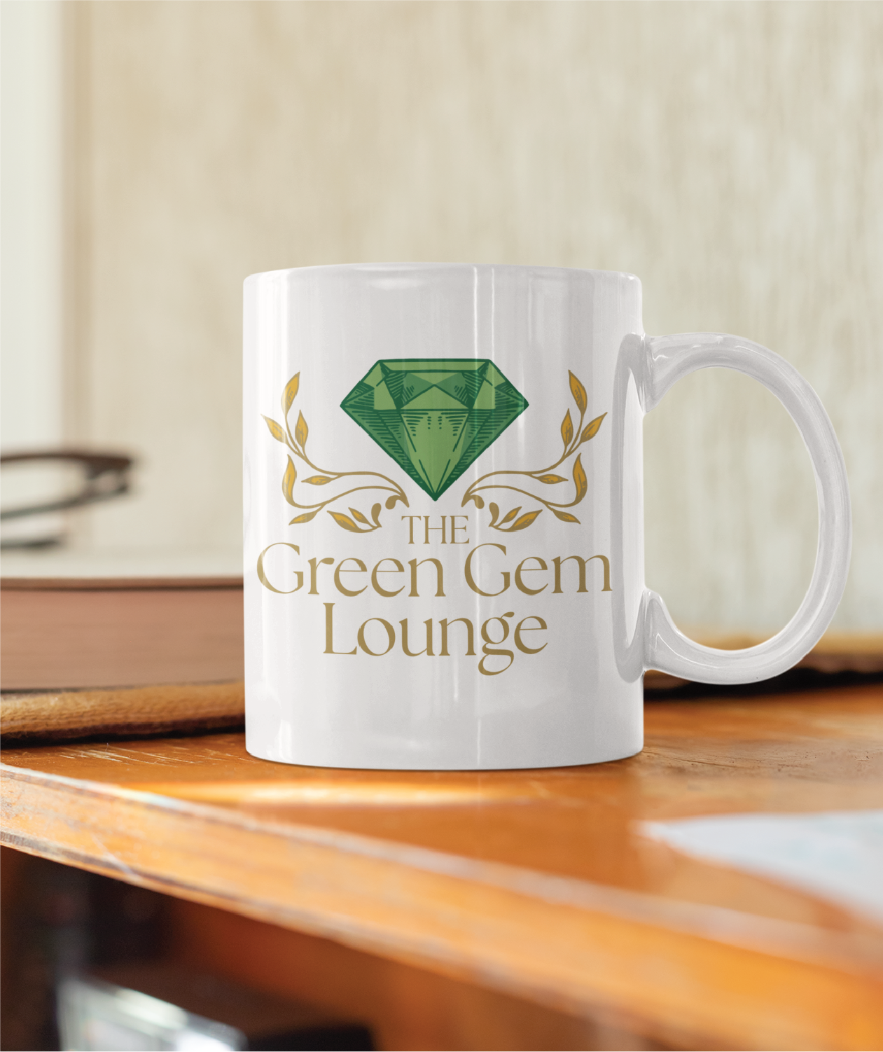 A white mug on a wooden surface displays a large green gemstone, gold leaves, and the text The Green Gem Lounge in gold lettering—a perfect logo design inspiration for any Pennsylvania business. A blurred book and glasses are in the background.