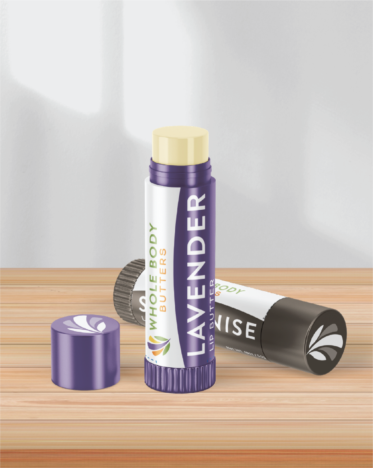 Two lip balm tubes on a wooden surface; one upright purple tube labeled Lavender Whole Body Butters, featuring a custom print design, and one black tube lying down with a leaf logo. Light shines on the background wall.