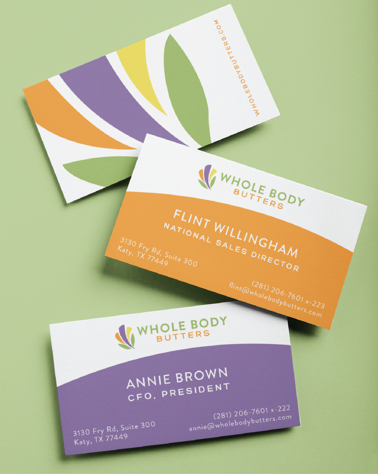 Three colorful business cards for Whole Body Butters are displayed on a green surface, showcasing custom print design. Each card features a leaf motif, brand logo, contact details, and titles for Flint Willingham and Annie Brown.