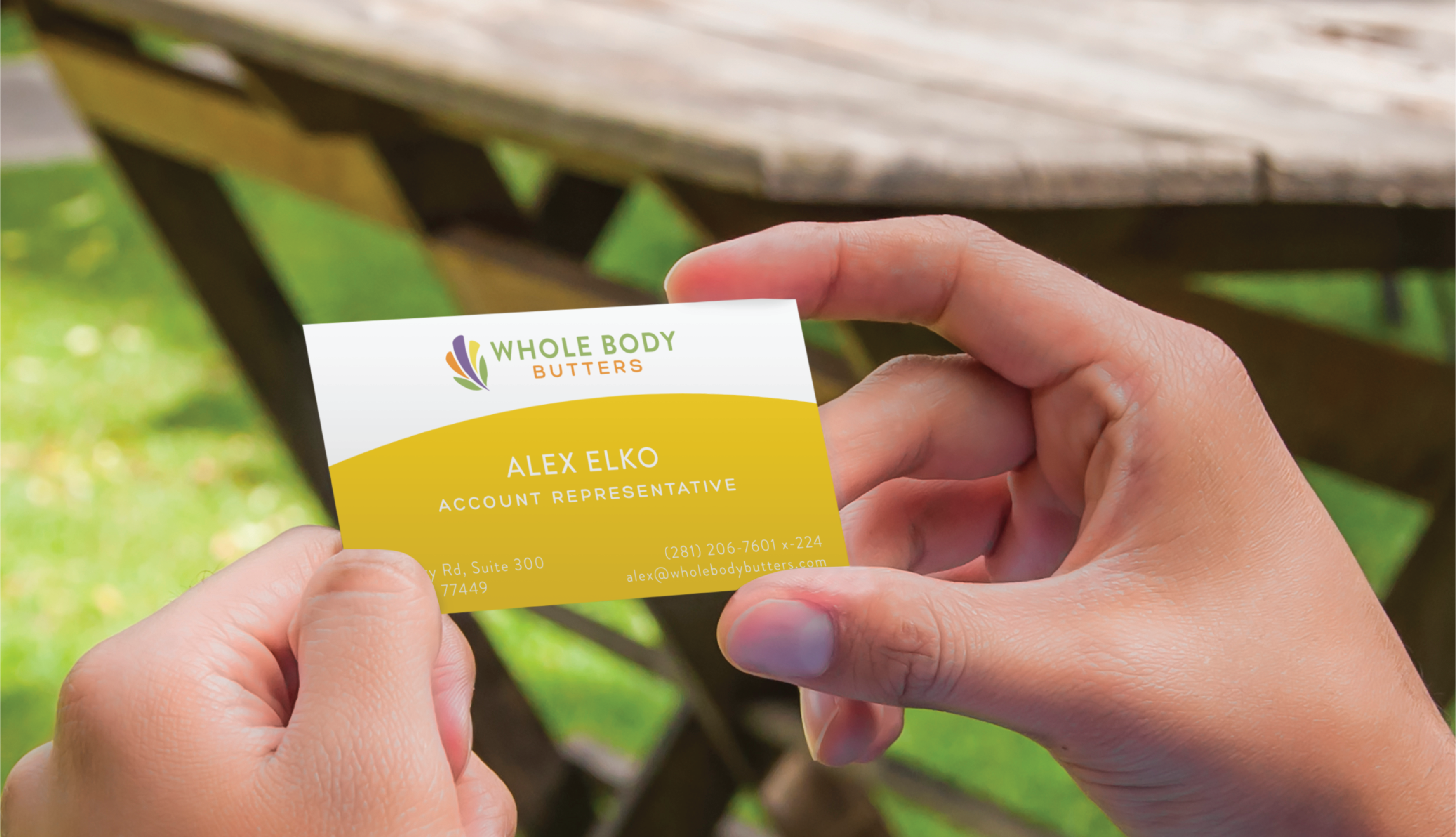 professional business card design | Moonlit Media LLC A person holds a business card for Whole Body Butters featuring Alex Elko, Account Representative. The custom print design stands out against a blurred wooden table and green outdoor scenery in the background.