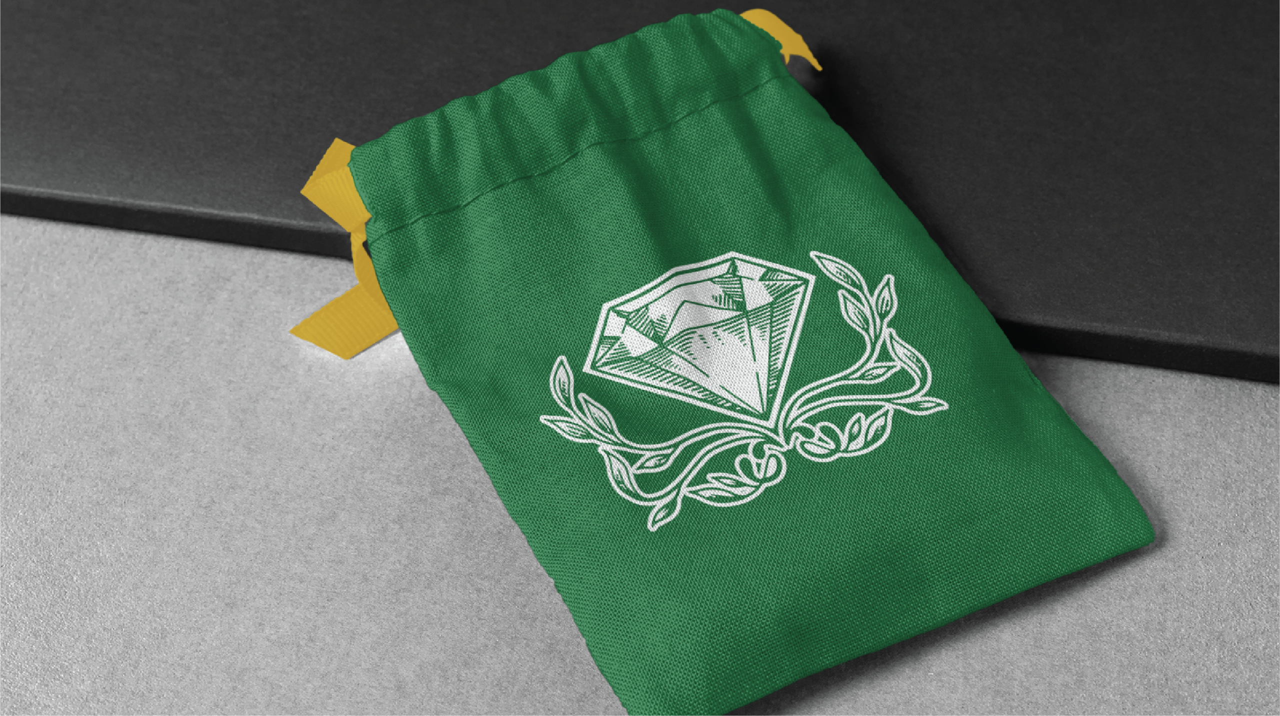 A green fabric drawstring pouch with yellow ribbons, featuring a white diamond and laurel branch logo design on the front, lies on a gray and black surface.