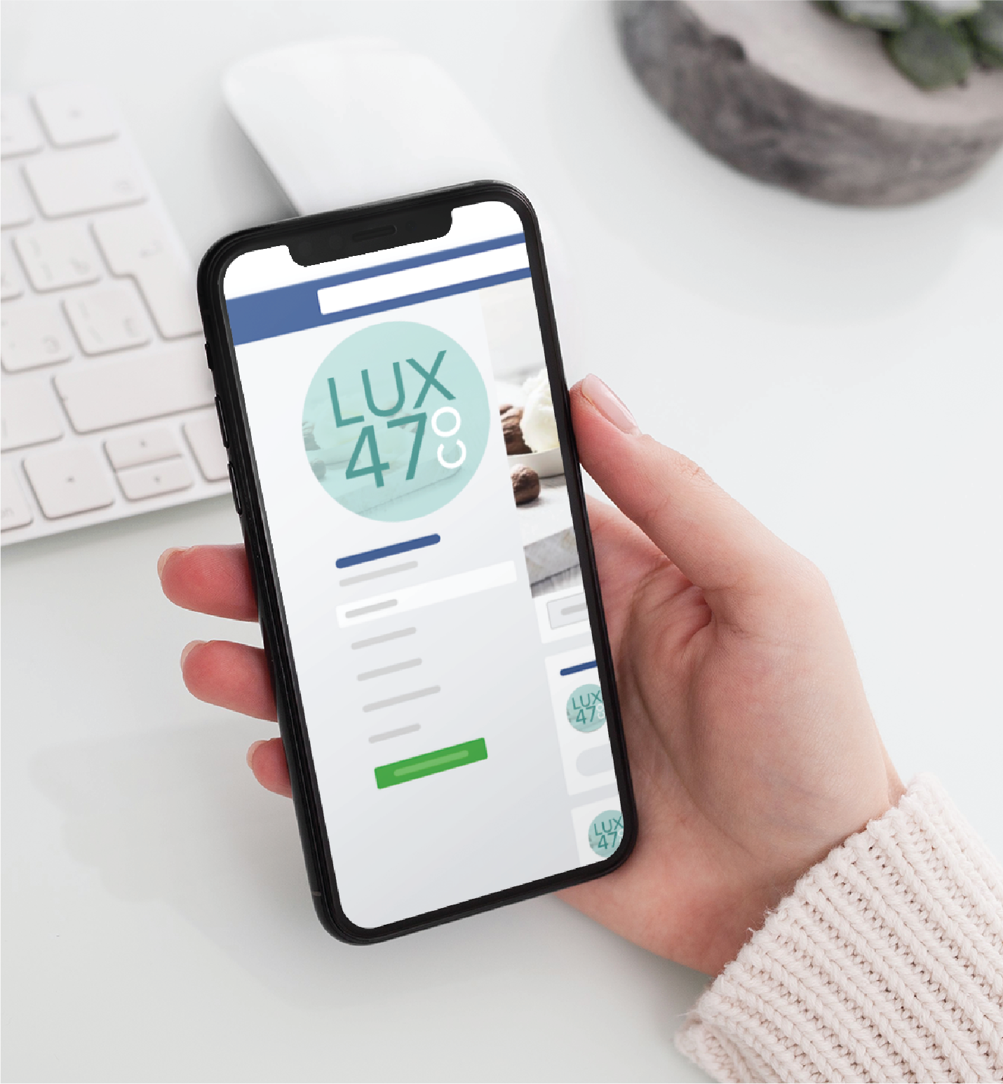 A person holds a smartphone displaying a webpage for LUX 47 CO. at a white desk with a keyboard, mouse, and plant in the background—perfect inspiration for Social Graphics for Realtors looking to enhance their online presence.
