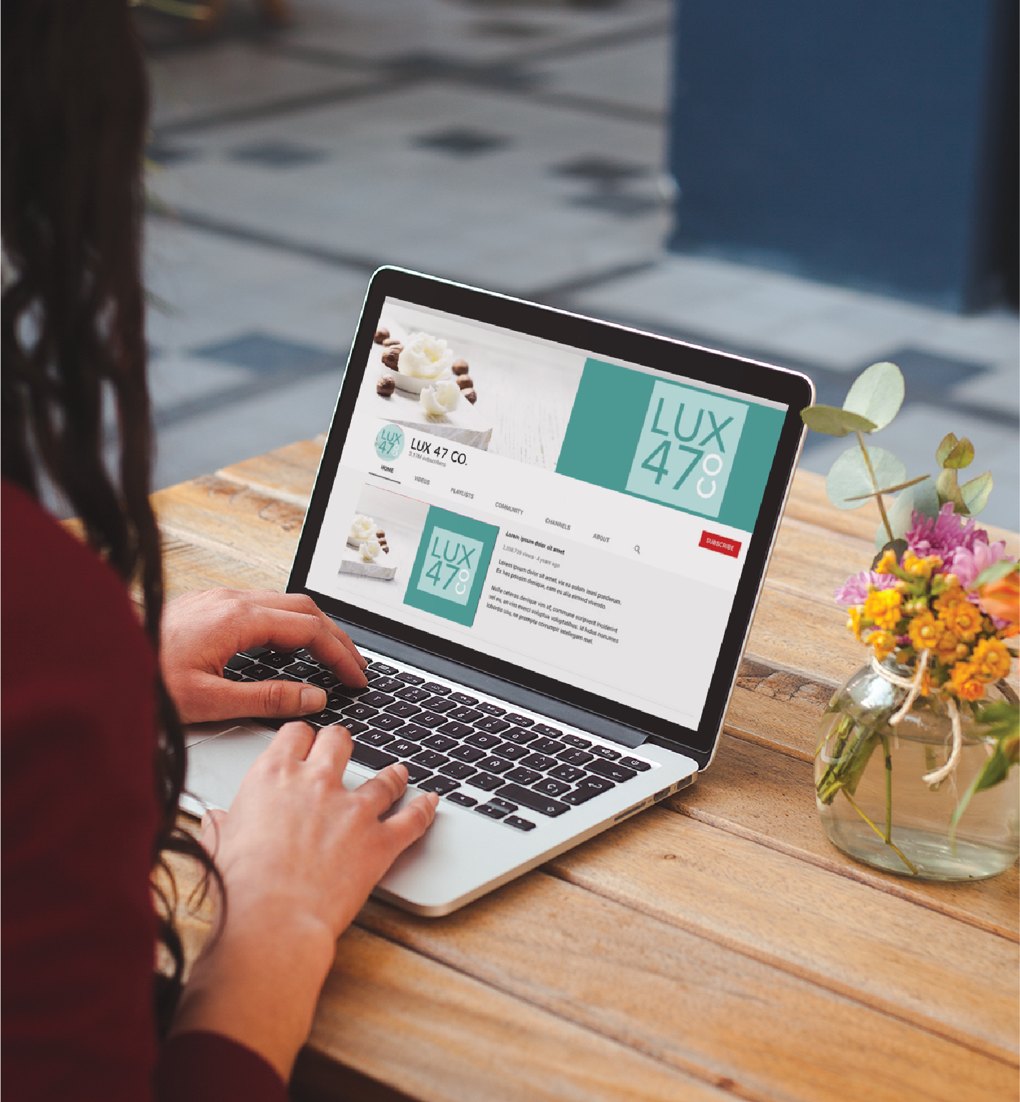 A person types on a laptop at a wooden table with a small flower vase. The screen displays the “LUX 47% CO.” YouTube channel, featuring teal branding, chocolates, and social graphics tailored for California realtors.