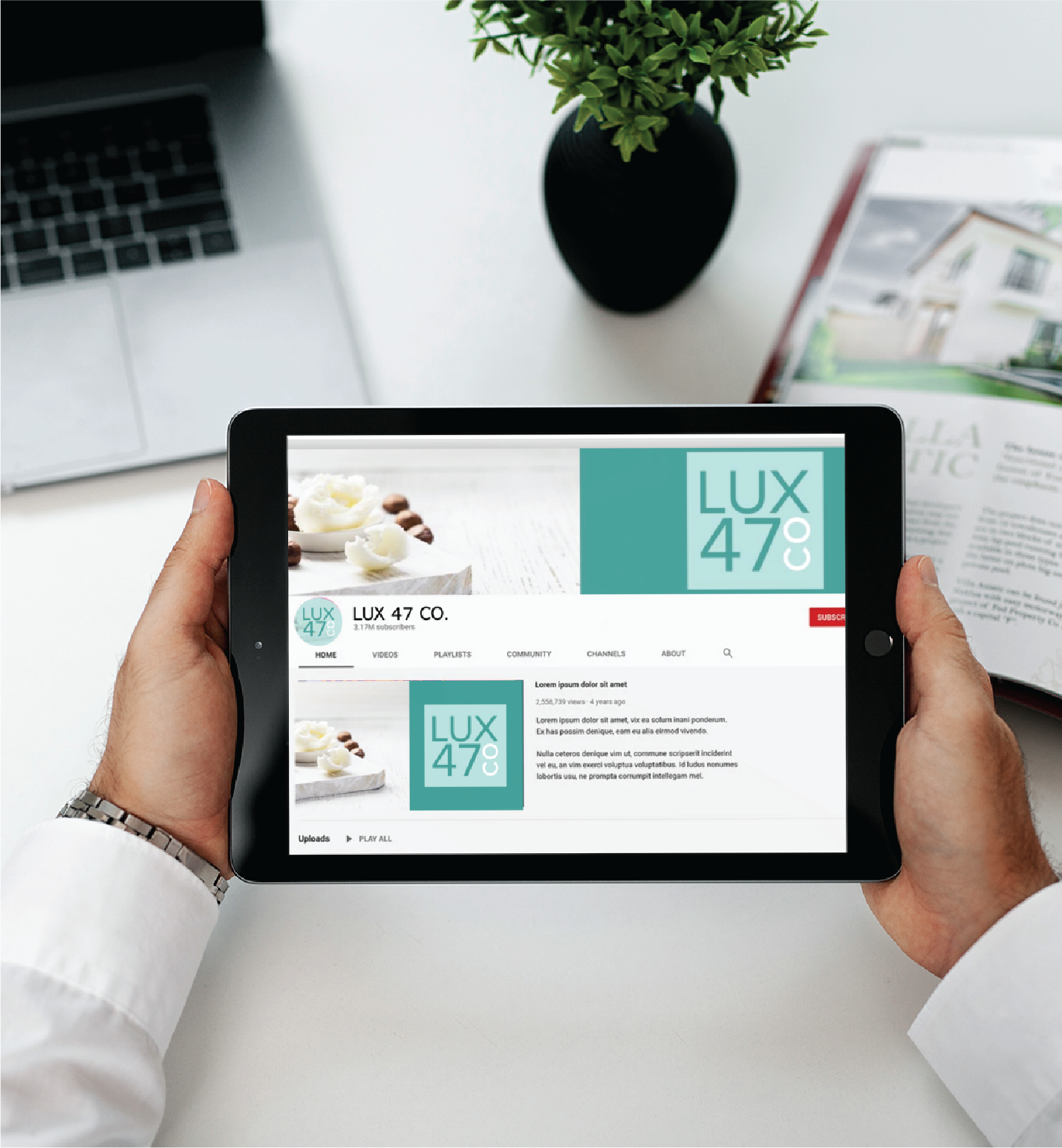 A person holds a tablet displaying the YouTube channel page for LUX 47 CO., a California-based Realtors group, at a desk with a laptop, plant, and open magazine. The channel logo and information are visible on the tablet screen.