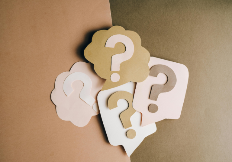 Paper cutouts in neutral tones shaped like speech bubbles and clouds, each featuring a large question mark, are arranged on a two-toned beige and brown background—perfect for illustrating FAQs on your small business website.