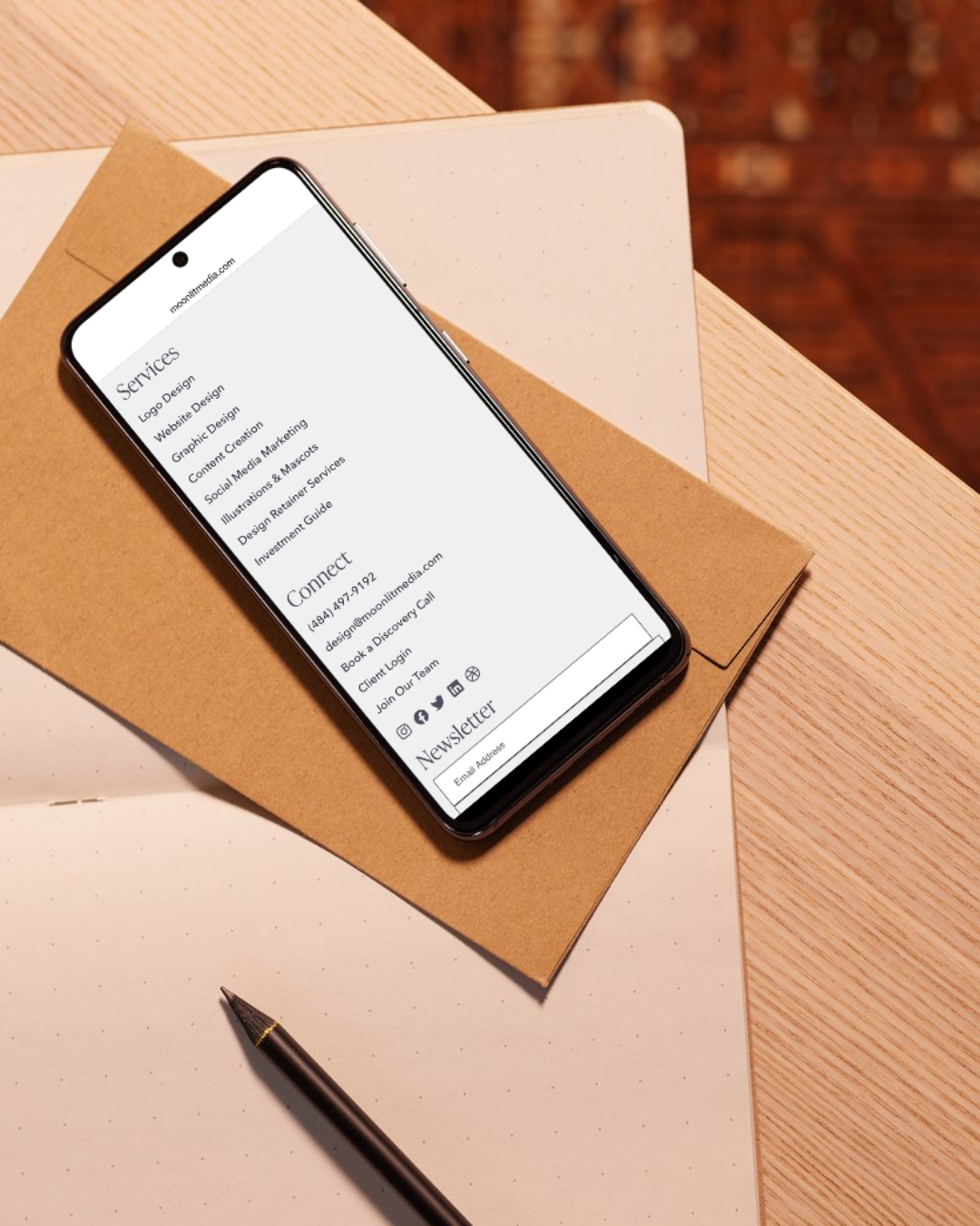 A smartphone displays a website’s services and contact info, including an improved website footer for easy access to customer resources, resting on a brown envelope atop a dotted notebook on a wooden desk. A black pen lies nearby.