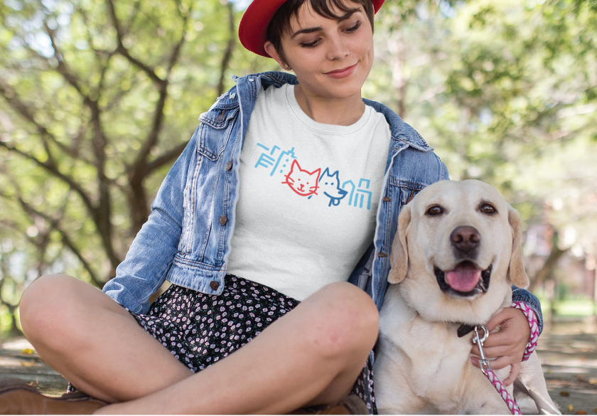 A woman in a denim jacket and red hat sits on a bench outdoors, smiling down at a yellow Labrador Retriever beside her. Her T-shirt, printed with a cute cat graphic featuring blue lines and a red outline, looks like something you’d find from creative local businesses that specialize in unique print marketing.